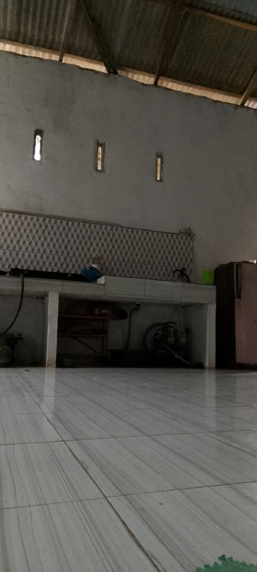 The image shows an outdoor covered area with a corrugated metal roof supported by wooden beams. The walls are light gray, and three small, rectangular openings are visible near the top of the wall. Below the openings, a tiled backsplash with a diamond pattern runs across the wall. A countertop extends horizontally, supported by white bases. On the countertop, a blue cloth object is present. Below the countertop, a red shelf unit is visible. Various items are located under the countertop, including what appears to be a motor or engine with wires and a green container. To the right, a portion of a light brown refrigerator is visible. The floor is tiled with light gray tiles that have a wood-grain pattern. The scene appears to be in Karang Gading, Indonesia, likely an outdoor kitchen or service area.