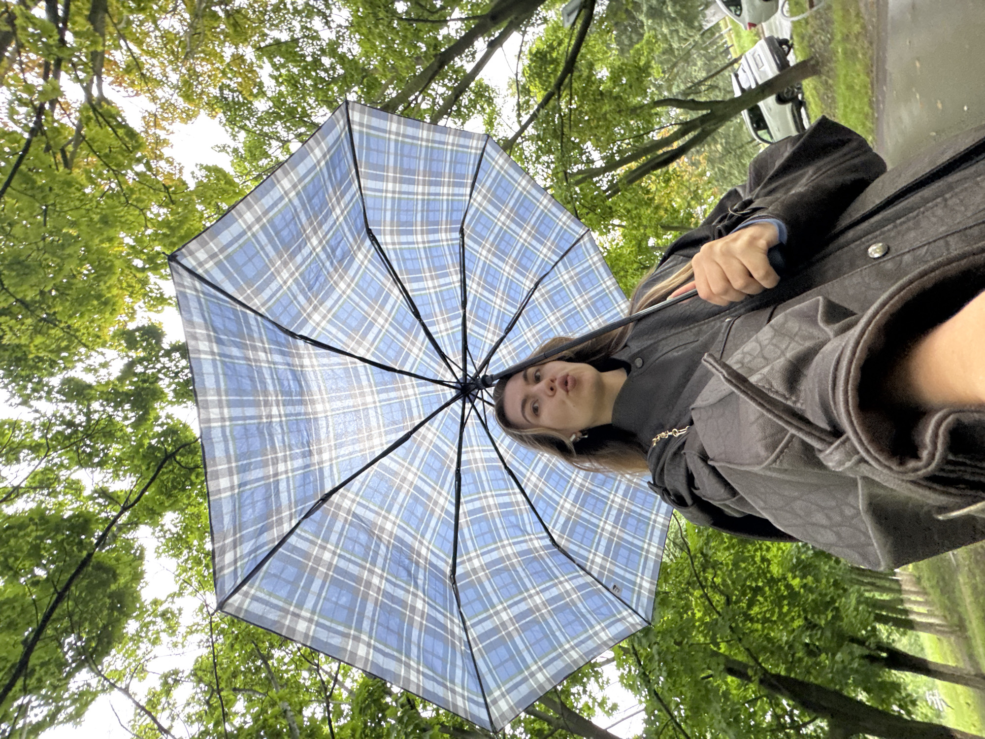 An outdoor, low-angle, medium shot captures a young woman standing on a sidewalk, holding a large, blue and white plaid umbrella open above her. She is facing the camera, her mouth puckered, and her hair is brown and long. She wears a dark coat with a textured pattern and a gold chain strap purse on her shoulder. The background is a park-like setting with lush green trees on either side of a wet asphalt road. Several cars are parked along the side of the road, and the sky is overcast. The overall impression is one of a cool, damp day in an urban park.