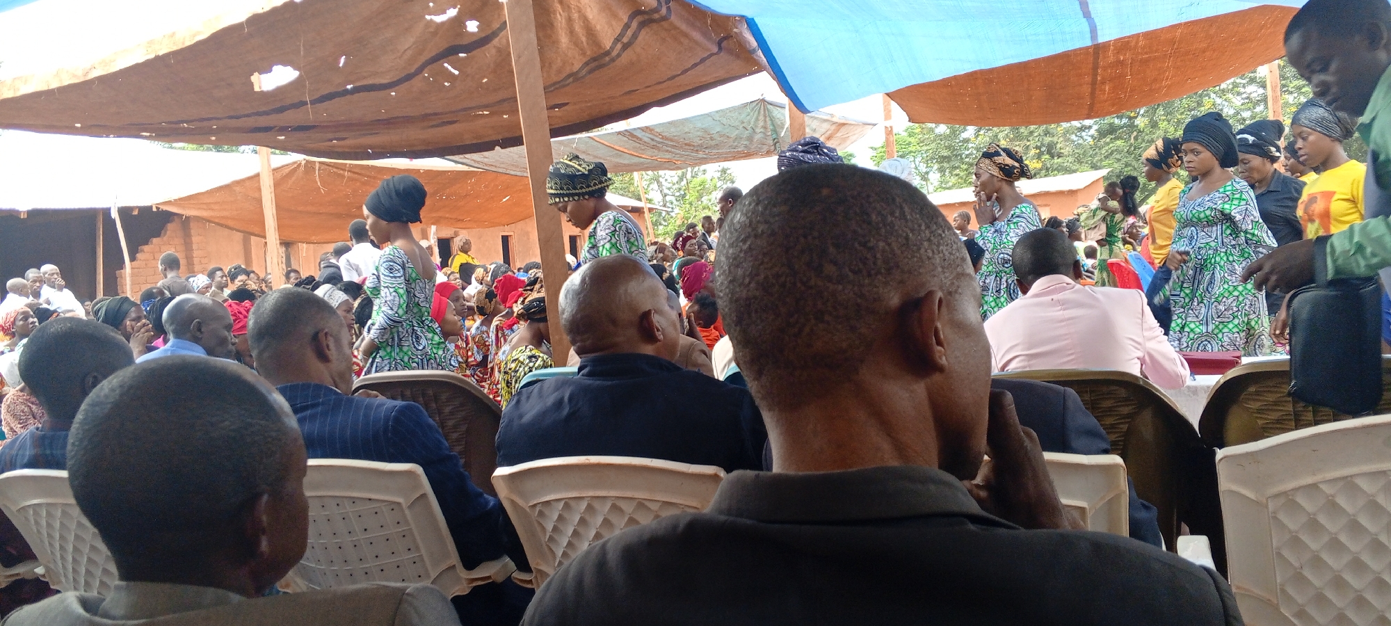 A gathering of people is occurring in Ngalula, Democratic Republic of the Congo. Several individuals sit in white plastic chairs beneath a makeshift canopy of light brown and blue tarps. The crowd includes men with shaved heads and women wearing vibrant patterned dresses and headwraps. Some attendees stand along the perimeter. The atmosphere appears to be that of a formal or semi-formal event.