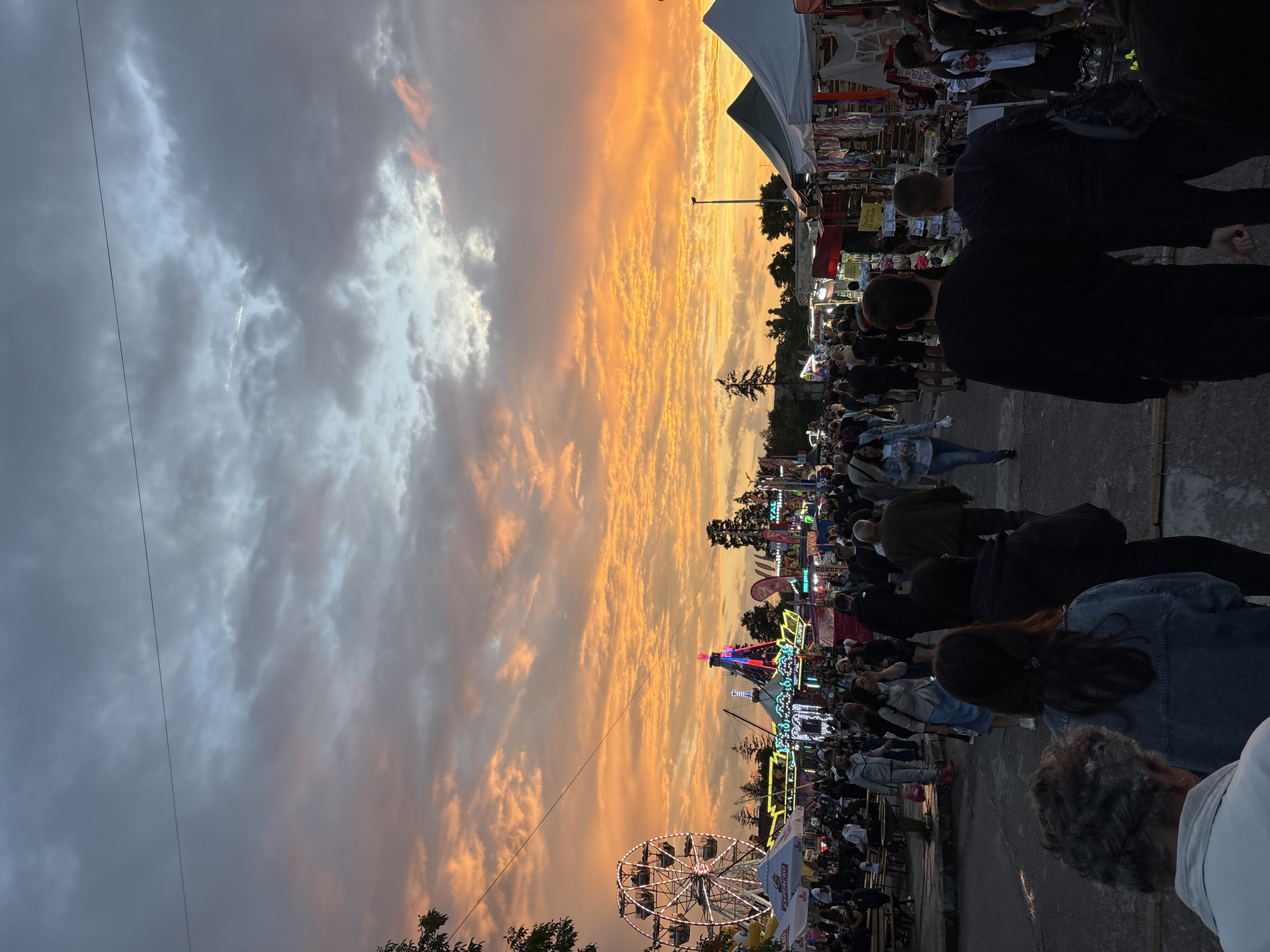 The image depicts a crowded outdoor fair or market scene in the city of Elin Pelin, Bulgaria, during sunset or dusk. The sky occupies the upper two-fifths of the frame, transitioning from a darker, overcast grey-blue at the top, horizontally bisected by a thin dark wire, to a vibrant orange and red expanse of altocumulus clouds in the mid-sky, indicating a low sun angle.
Below the sky, the midground is dominated by various fair attractions and numerous people. On the far left, a large Ferris wheel with white illuminations is partially visible, obscured by dark tree foliage. Adjacent to it are white canopies or tents, one displaying Cyrillic text, suggesting food or beverage vendors. Towards the center and right, multiple brightly lit fair rides with multi-colored lights (red, blue, yellow) are active, including what appears to be a carousel and other spinning attractions. Further to the right, a series of vendor stalls with green and white pitched roofs line a path, displaying various goods. Dark silhouettes of trees and utility poles punctuate the horizon against the vibrant sky.
The foreground is filled with a dense crowd of people walking along a dark asphalt or concrete pathway. Most individuals are seen from behind or in profile, moving generally towards the background attractions. Their attire is casual, including dark jackets, denim, and light-colored tops. Specific individuals in the immediate foreground include a person with grey, curly hair wearing a light top (bottom left), another with long, dark hair wearing a denim jacket (center), and two males in dark clothing (bottom right). The ground appears dark, with some faint reflections of ambient light. The scene indicates a lively evening gathering, with artificial lights beginning to dominate as natural light fades.