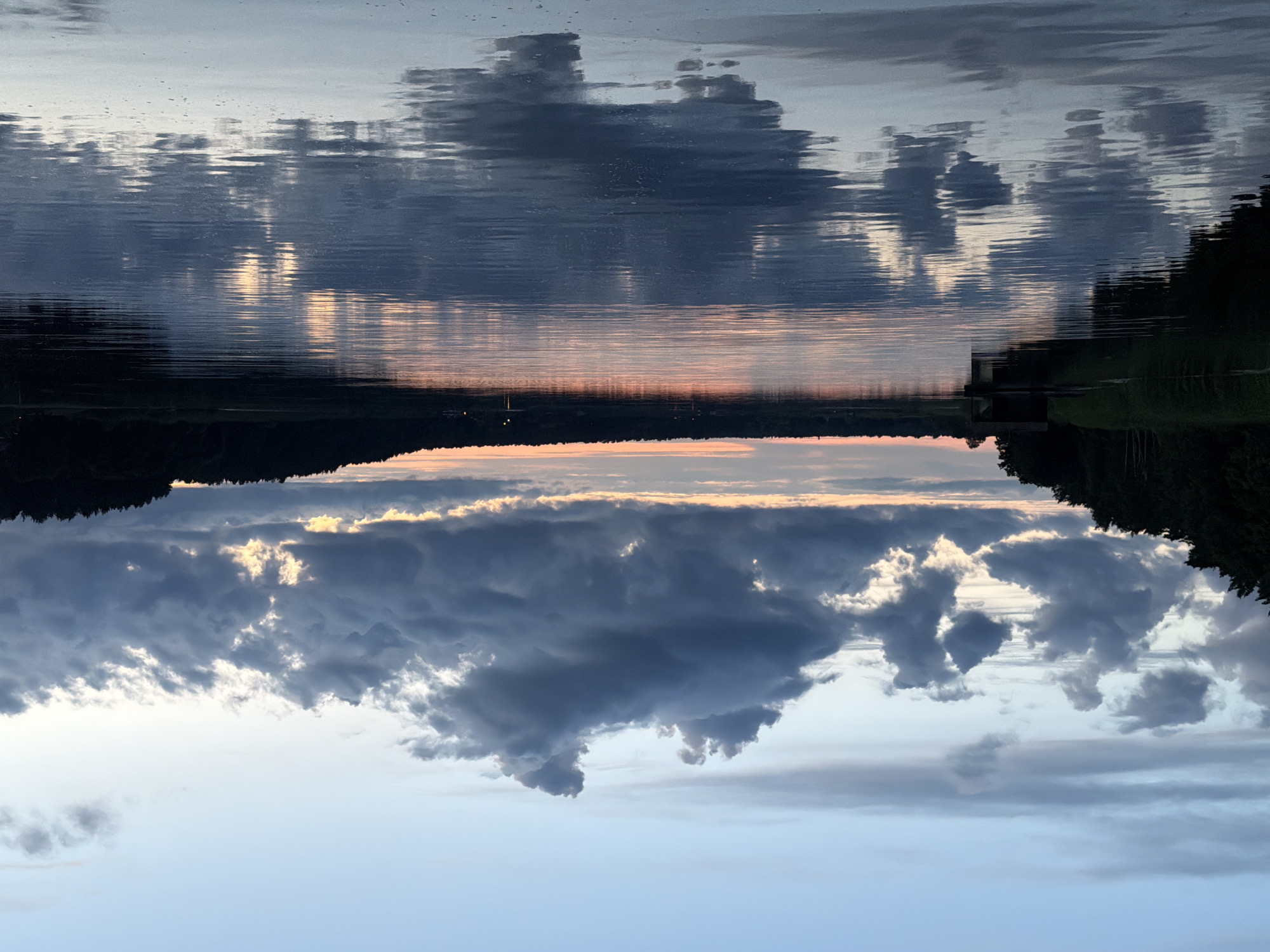 The image depicts a tranquil lake scene at dusk in Varkaļi, Latvia. The sky is a gradient of soft blues and grays, with dramatic, dark clouds scattered across the upper half, interspersed with patches of pale orange and yellow light near the horizon, indicative of the setting sun. The clouds cast reflections on the calm surface of the lake, which occupies the lower two-thirds of the frame. The water is smooth, with subtle ripples that distort the reflections of the sky and the dark silhouette of a tree-lined shore on the far side. On the left side of the frame, a dark wooden structure, possibly a boathouse or a small pier, is visible near the water's edge, partially obscured by reeds. The overall atmosphere is serene and peaceful, characterized by the muted colors and the quiet stillness of the natural environment.