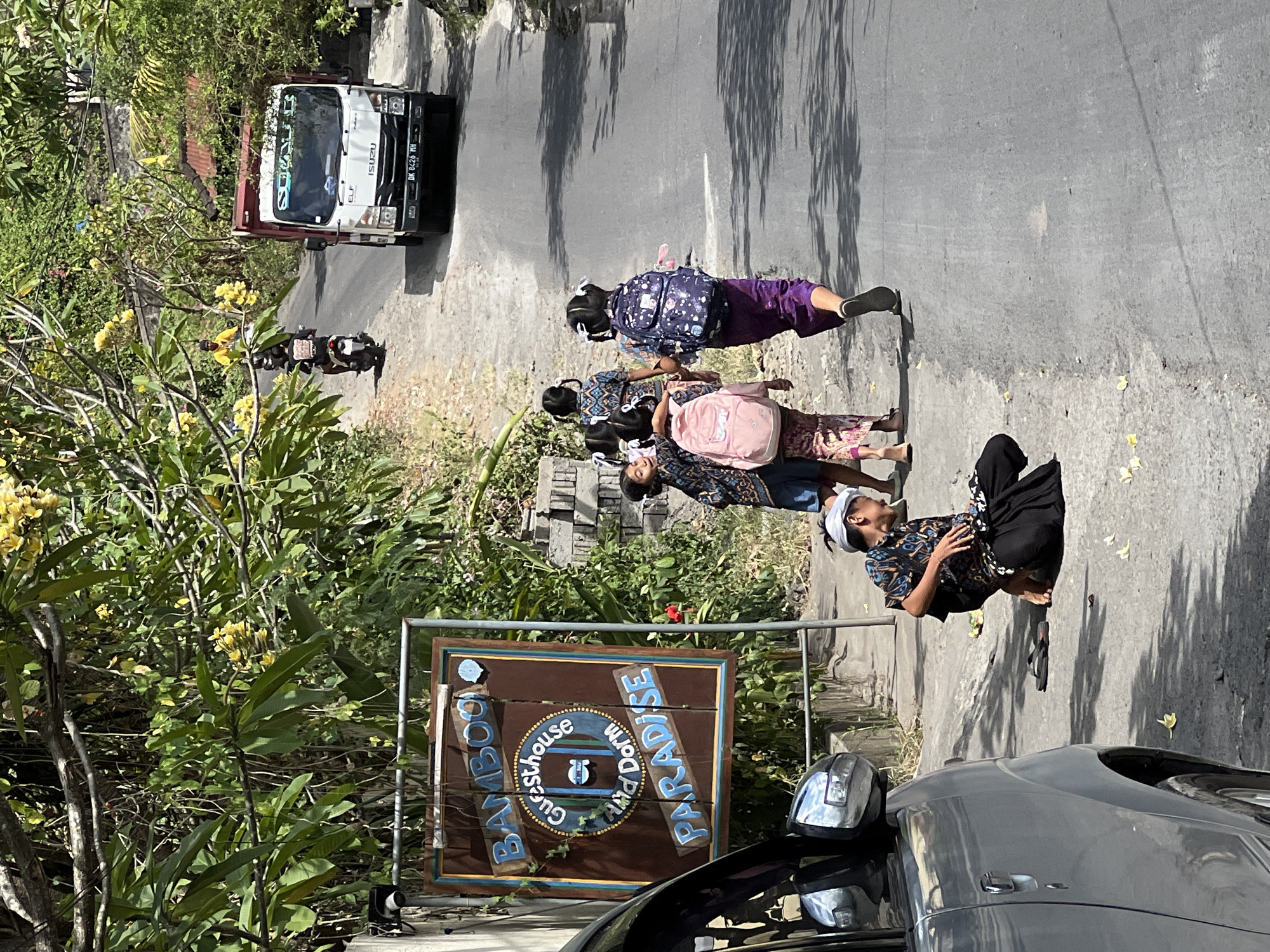 In Manggis, Indonesia, a street scene unfolds under a bright, sunny sky. A silver car is parked on the left side of the asphalt road. A sign for "BAMBOO Guesthouse and Dorm PARADISE" stands near the car. In the middle ground, a young boy in traditional clothing is kneeling on the side of the road. Further down the road, a group of four young girls, some carrying backpacks, are walking away from the viewer. To the right, a white and red ISUZU truck is driving down the road. The environment is lush and green, with trees and foliage lining the sides of the road.