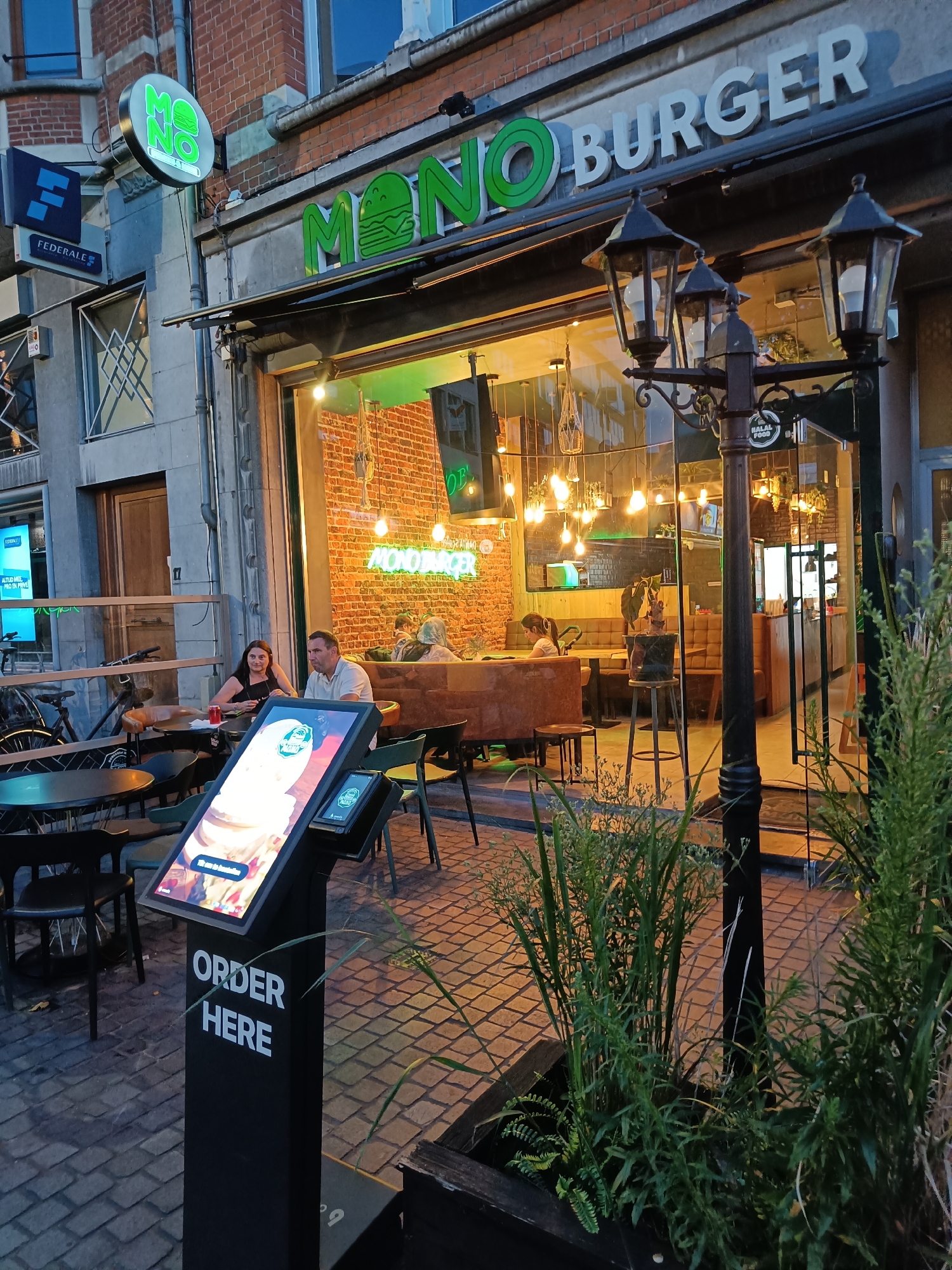 The image shows a street-level view of a restaurant called "MONO BURGER" in Leuven, Belgium. The restaurant has a large glass facade revealing an interior with brick walls, booth seating, and various light fixtures. Outside, there are tables and chairs set up on a cobblestone patio. A prominent digital ordering kiosk is positioned in the foreground, displaying the text "ORDER HERE" on its base and an advertisement on its screen. Two people are seated at a table on the patio, a woman on the left and a man on the right. Several other patrons are visible inside the restaurant. A street lamp with three lights stands near the entrance. The signage above the restaurant reads "MONO BURGER" in prominent green lettering. A smaller circular sign with the "MONO" logo is mounted to the left of the main sign. A blue sign with a white "F" logo and the text "FEDERALE" is visible on the building to the left of the restaurant. Green plants are in a planter in the lower right corner of the image. The lighting suggests it is evening or dusk.