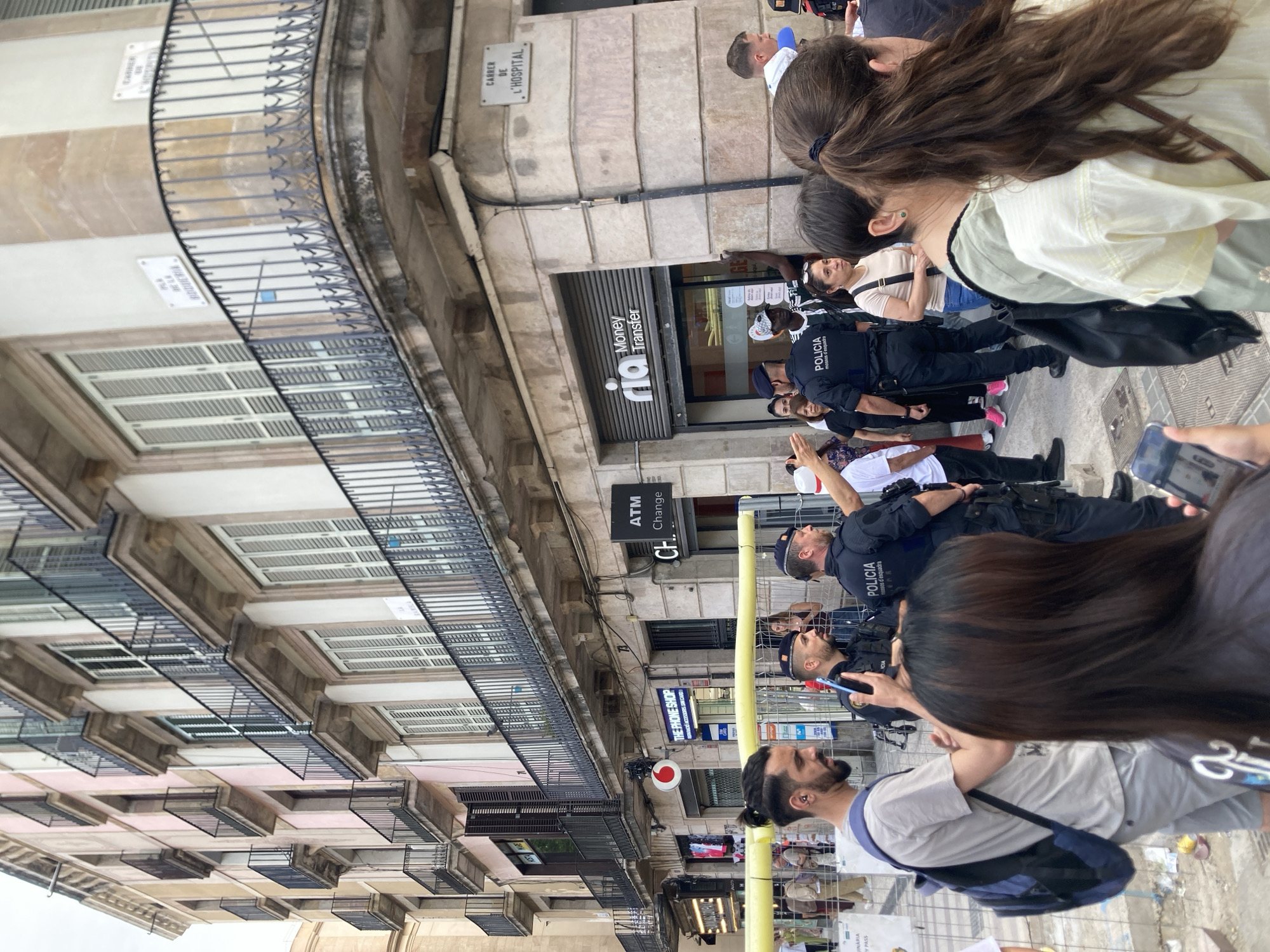 This image depicts a street scene in Barcelona, Spain, likely during the daytime. The focus is on a group of people, including several police officers in uniform, standing on a paved street. The officers are wearing black uniforms with "POLICIA" and "mossos d'esquadra" visible on their vests, indicating they are part of the Catalan police force. Some officers are holding mobile phones.
The setting is an urban environment with buildings lining the street. A prominent feature is a building with a balcony railing and balconies with slatted windows. Several signs are visible on the buildings, including one for "Money Transfer" with the "rio" logo, an "ATM Change" sign, and a sign for "THE PHONE SHOP" selling accessories and SIM cards. There's also a street sign that reads "CARRER DE L'HOSPITAL," confirming the location in Barcelona.
The people in the image appear to be a mix of civilians and police officers, suggesting a possible event or gathering is taking place, though the specific nature of the activity is not immediately clear. Some individuals are looking towards the camera or in the direction of the police. The overall atmosphere seems neutral, with no overt signs of distress or celebration. The weather appears to be overcast or hazy.