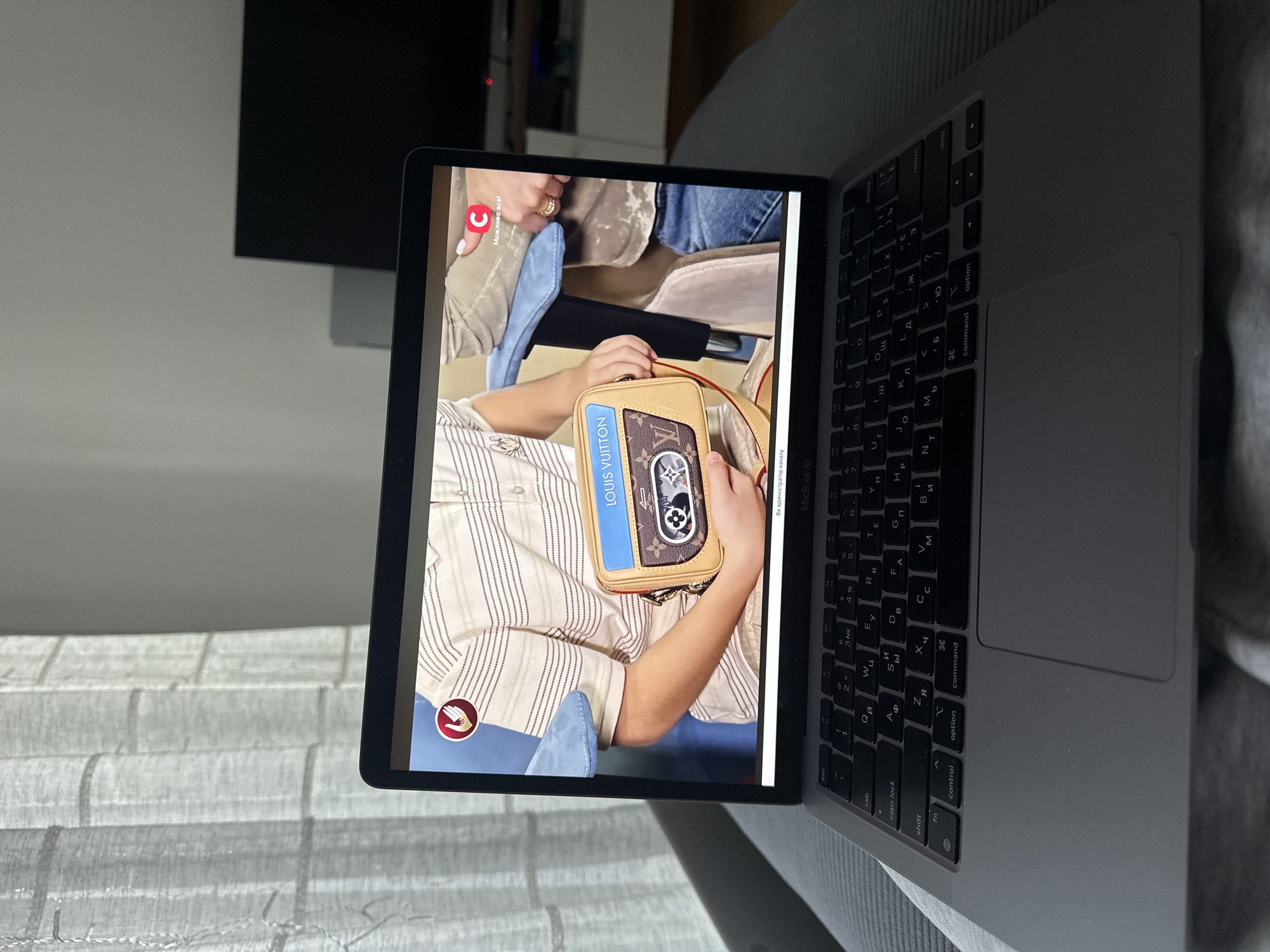 The image shows a gray laptop displaying a video of a person holding a Louis Vuitton handbag. The laptop is positioned on a gray soft surface. In the background, there is a window with white curtains, a white cabinet, and a black TV screen. The scene is indoors. There are no identifiable people or actions besides the laptop displaying the video. The specific location in Donji Pobori, Montenegro, cannot be determined from this image.
