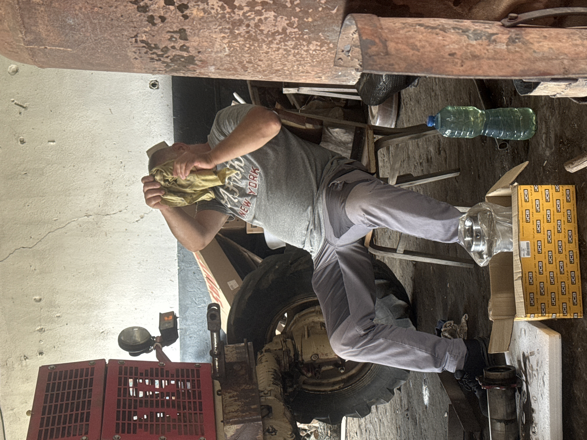 This image captures a man engaged in work within what appears to be a rustic workshop or garage, likely in Sarnevo, Bulgaria. The central subject is a man, seen from the side, wearing a grey t-shirt with "NEW YORK" printed on it and grey trousers. He is bent over, holding a yellowish cloth to his face or head, possibly wiping sweat or dirt from his brow while working on heavy machinery.
He is positioned next to a large tire and other components of what looks like a tractor or similar industrial vehicle, indicated by visible red grates and a headlight on the left side of the frame. Nearby, on the floor, are several cardboard boxes. One prominent box, opened, contains new parts, and displays the "JCB" brand name repeatedly, along with some Arabic script. A green plastic bottle with liquid stands upright on the floor next to other miscellaneous items and a white styrofoam block.
The environment is industrial and somewhat cluttered, characterized by a rusty metal structure overhead, a light-colored concrete wall with some cracks, and a dirt or concrete floor. The lighting suggests an indoor setting, possibly with natural light entering from an unseen source. The overall scene depicts a moment during manual labor or mechanical repair in a practical, functional workspace.