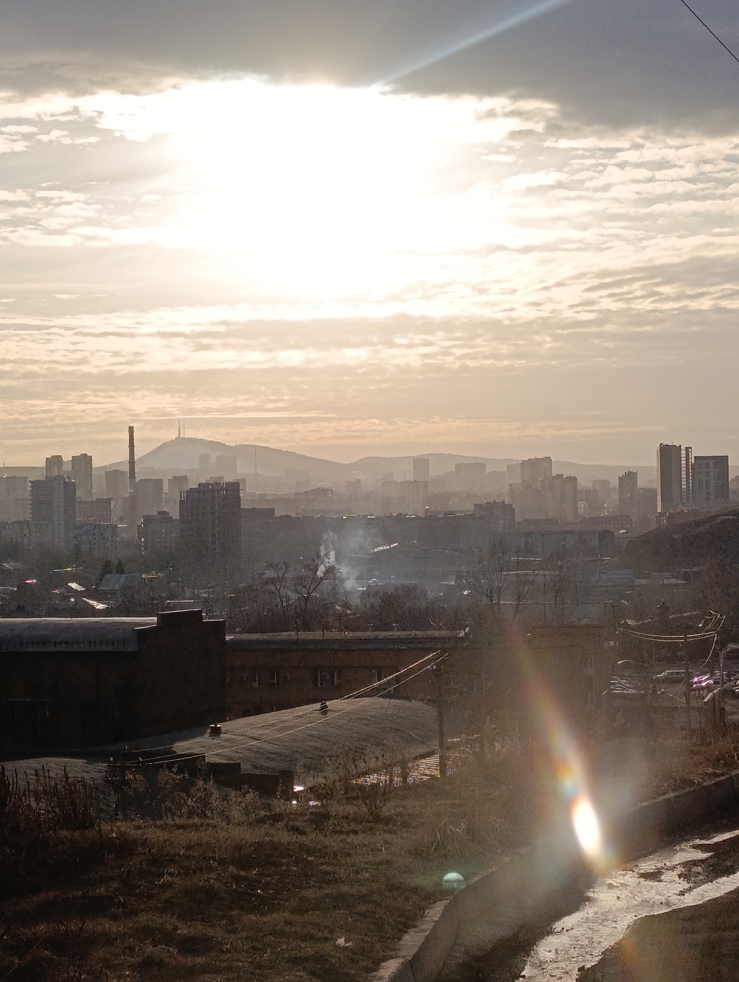 The image displays a panoramic urban landscape of Krasnoyarsk, Russia, observed from an elevated vantage point. The scene is illuminated by strong, low-angle sunlight from the upper right, causing significant lens flare that manifests as a diagonal streak and a bright oval shape in the lower right foreground.
The sky occupies the upper portion, characterized by bright, hazy sunlight partially obscured by scattered cumulus clouds. The atmospheric conditions suggest a diffuse light, possibly indicative of early morning or late afternoon.
In the distant background, a sprawling cityscape extends to a range of hills. A prominent, conical mountain, identifiable as Karaulnaya Mountain due to the distinct communication tower visible on its summit, anchors the horizon. A tall, slender industrial chimney, characteristic of the city's power infrastructure, rises significantly above the other buildings to the left of the mountain. The distant urban area is dense with multi-story residential and commercial buildings, appearing partially obscured by atmospheric haze. Small plumes of white smoke are visible rising from multiple points within the city's midground.
The midground features numerous dark-colored, multi-story buildings, consistent with older urban or industrial architecture. Sparse, bare deciduous trees and shrubs are interspersed among these structures, indicating a season devoid of foliage, such as late autumn or winter.
The immediate foreground consists of a gently sloping earthen area covered with dry, brownish vegetation, primarily dormant grasses and sparse undergrowth. To the right, a concrete channel or pathway, partially covered with patches of white material resembling snow or ice, descends out of frame. No individuals are discernible within the frame.