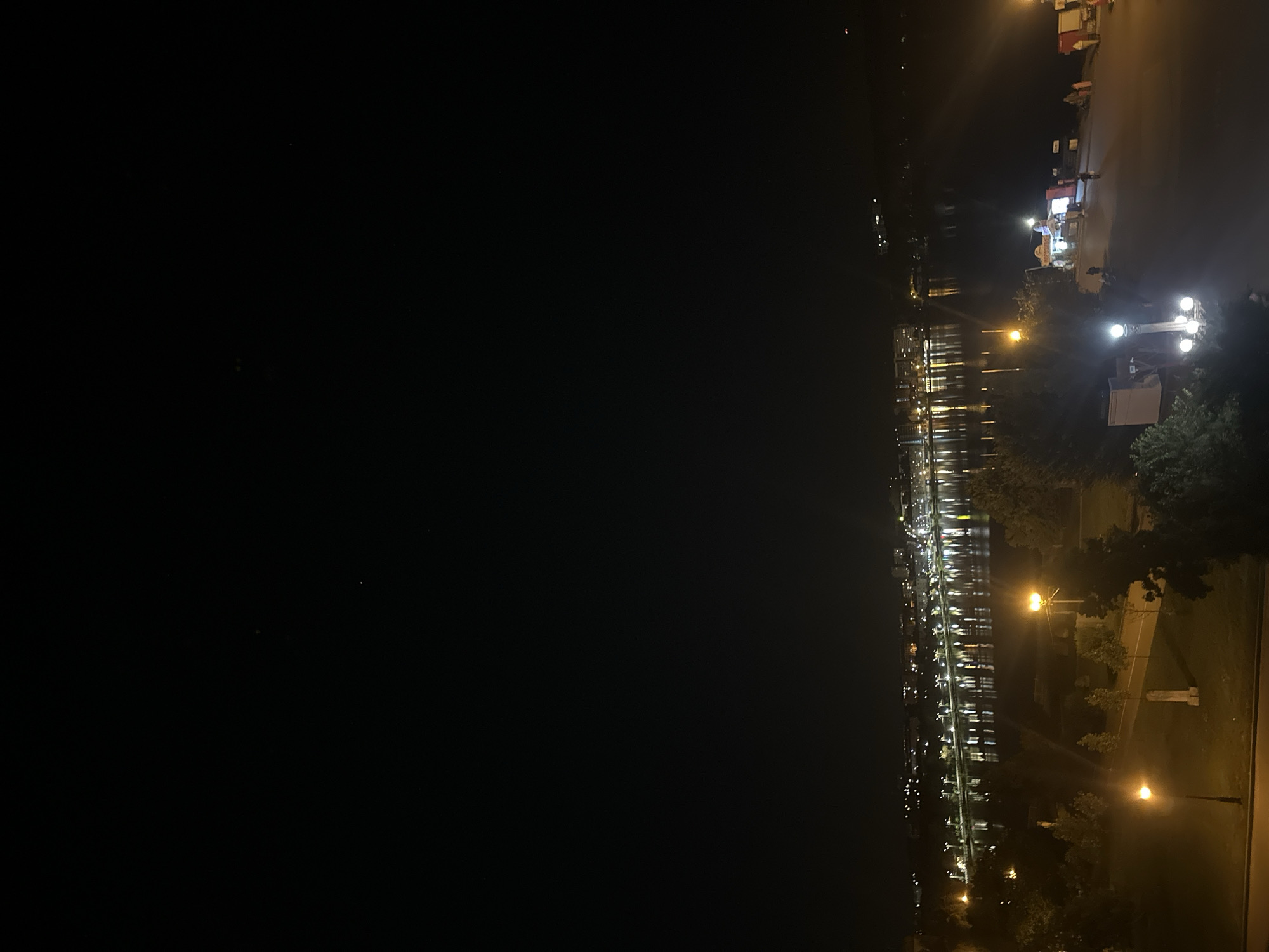 The image depicts a nighttime view of a city park and waterfront, likely in Ternopil, Ukraine, overlooking a body of water with a city skyline illuminated in the distance. The foreground features a park area with trees, grassy patches, and walking paths. Several street lamps are visible, casting a warm yellow light on the scene. One prominent lamppost with multiple bulbs stands out. There are a few structures that appear to be kiosks or small stalls set up in the park area, with some lights on. A few people are visible in the park, walking or standing, though their details are indistinct due to the low light. The middle ground shows the expansive, dark body of water, reflecting the lights of the distant city. The city skyline across the water is lined with numerous lights from buildings and streetlights, creating a shimmering effect on the water's surface. The sky above is dark, with a few faint stars visible. The overall atmosphere is calm and serene, characteristic of a city at night.