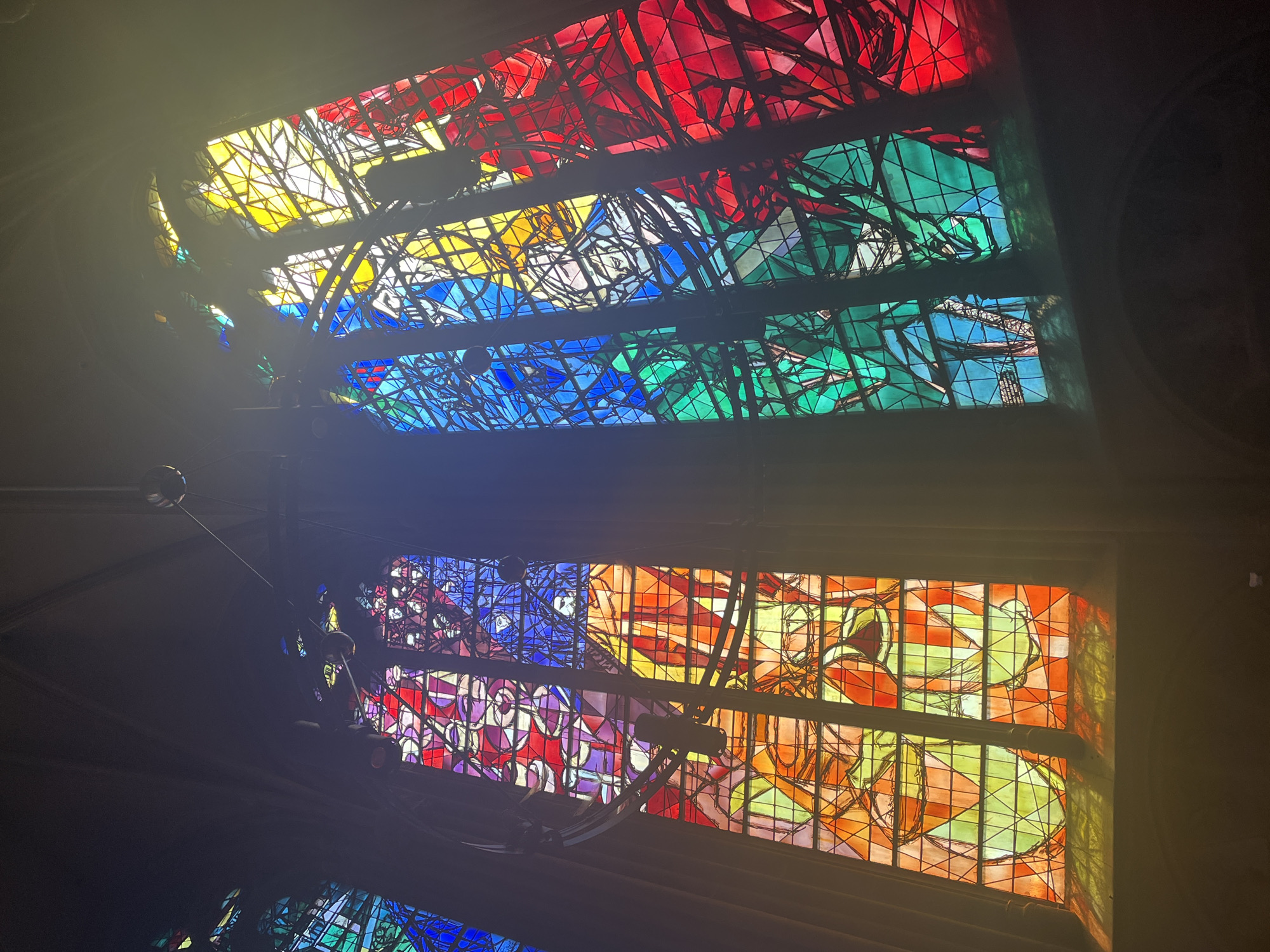 The image depicts a stained glass window inside the Saint-Étienne Cathedral in Metz, France. The window consists of colorful panes in red, yellow, blue, green, and orange hues separated by black lead lines. Abstract shapes and designs are visible within the panes. There are visible lights. The surrounding area is dark, suggesting an interior shot. Parts of what appear to be lighting fixtures and cables are visible against the window.