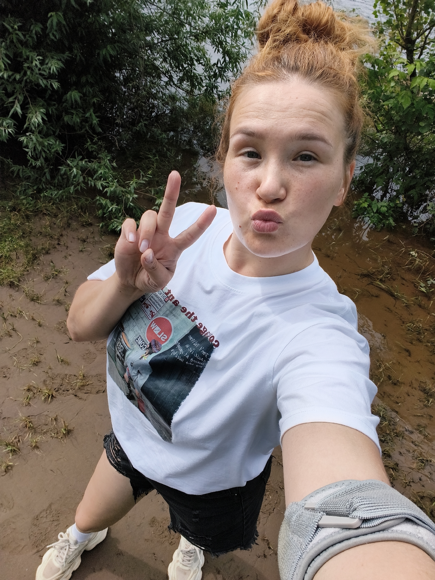 A young woman with light skin and auburn hair pulled up in a messy bun takes a selfie. She is wearing a white t-shirt with a graphic print, black distressed denim shorts, white socks, and white athletic shoes. Her left hand is raised, forming a peace sign, while her right arm is not visible. She is puckering her lips, as if for a kiss.
The background suggests an outdoor setting, possibly a park or a natural area near water. Lush green foliage is visible behind her, and the ground appears to be muddy with some dry grass. The water in the background is murky, suggesting it might be a river, lake, or flooded area. The lighting indicates it is daytime, and the overcast sky could imply cloudy weather. The overall mood appears playful and casual, with the woman posing for a fun photo in what seems to be a relaxed, natural environment.
The t-shirt has a graphic print with Japanese text and what appears to be an image of a person. There is also a circular logo with red and white colors.