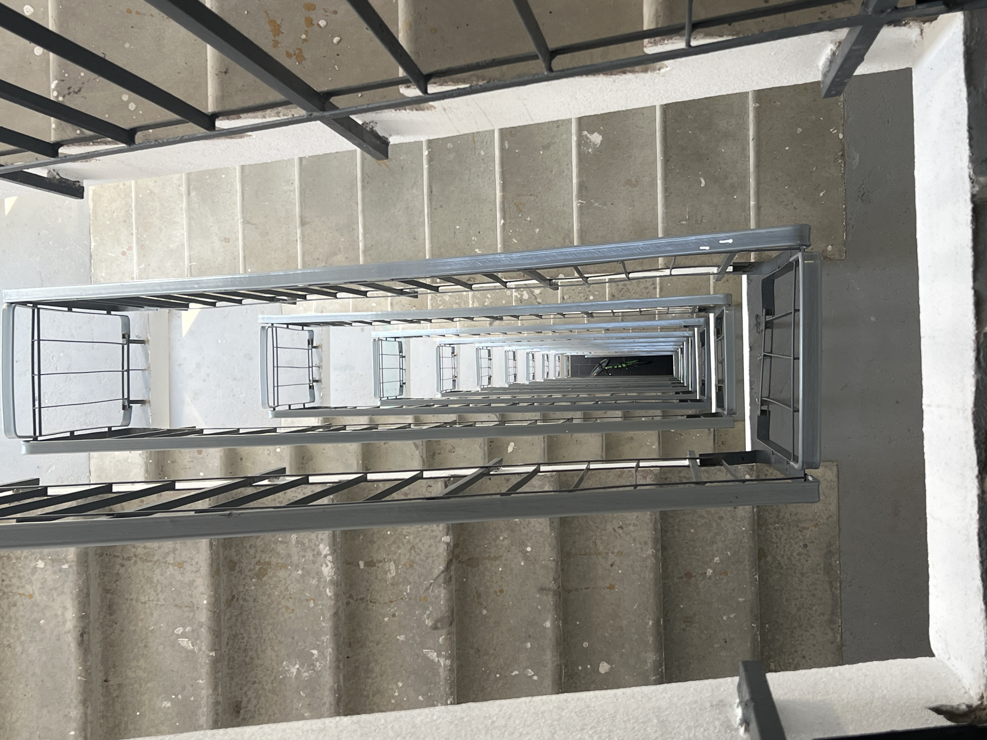 The image captures a high-angle view looking directly down a multi-story stairwell. The scene is dominated by parallel lines of concrete stairs and metal railings receding into the distance, creating a strong sense of depth.
The stairs are constructed from light grey concrete, exhibiting signs of wear, including scattered white and brown paint splatters, scuffs, and minor chips, particularly noticeable on the treads and risers. Two primary sets of stairs are visible, one descending on the left and another on the right, both spiraling downwards around a central void.
Parallel to each set of stairs are dark grey metal railings. These railings feature a rectangular top handrail supported by multiple vertical metal bars. The railings on the immediate left and right are sharply in focus, while those further down the stairwell become progressively smaller and less distinct due to perspective. The rightmost railing exhibits white scuff marks and some darker material adhering to its top bar.
The stairwell walls are painted white, showing some textured surfaces and minor imperfections. Several landings, flat sections of concrete floor, are visible at various intervals, providing a break between the descending flights of stairs. The floor on these landings appears to be a slightly darker shade of grey than the steps.
No individuals or animated subjects are present in the image. The lighting is even and diffuse, suggesting an interior space with overhead or window light from an unseen source. The stairwell terminates in a dark, indistinct area at the very bottom, implying many more unseen flights or a lower level.
Regarding the specific location in the city of Saint Petersburg, Russia, the image lacks any unique architectural features, signage, or contextual clues to forensically confirm this designation. The utilitarian design of the concrete stairwell and metal railings is common in various types of buildings, including residential, commercial, or industrial structures, across many different urban environments.