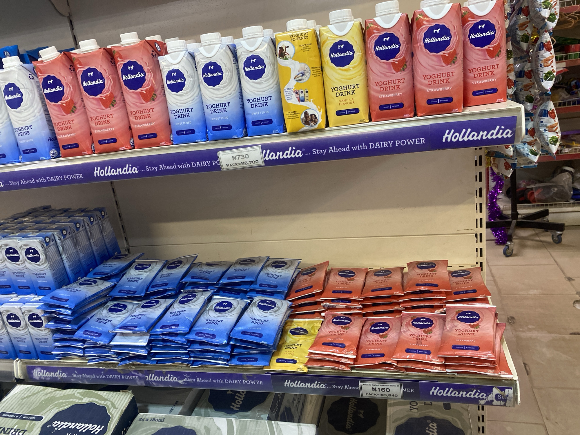 On a retail shelf in a store in Ikorodu, Nigeria, there are numerous cartons of Hollandia yoghurt drink. The cartons are arranged in rows and vary in flavor and size. Visible flavors include strawberry, vanilla, and a plain sweetened variety. Some cartons are larger upright containers, while others are smaller, flat pouches. The shelf itself has a purple trim with the Hollandia logo and slogan. In the background, other store items and fixtures are partially visible, including a mobile cart with wheels and decorative purple tinsel. The lighting is overhead and appears to be typical for a retail environment.