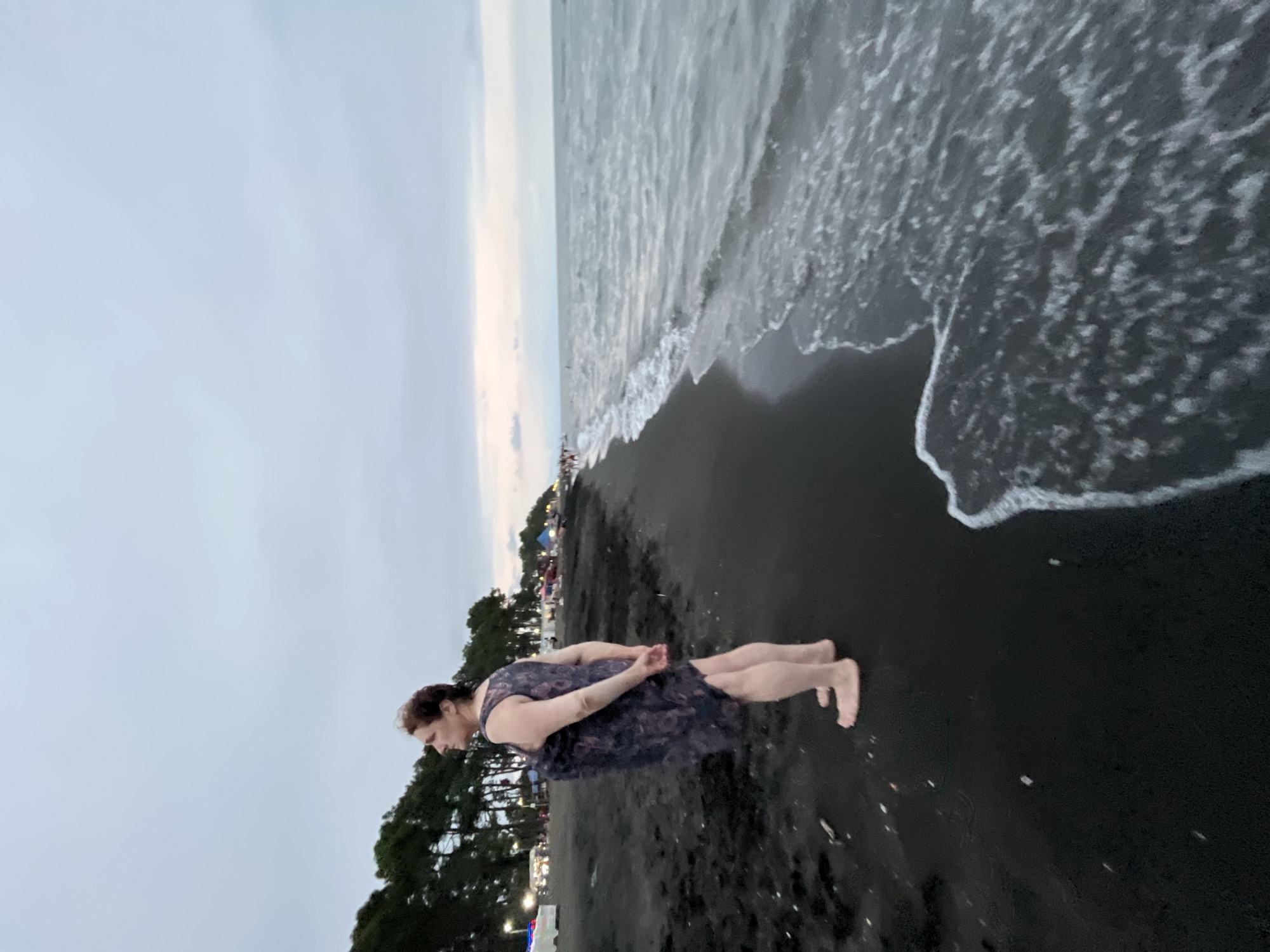 A woman stands on a black sand beach in Ureki, Georgia, looking out at the sea. She is wearing a dark, floral-patterned dress and is barefoot. Her hands are clasped behind her back. The sky is overcast, and the sea is choppy with waves breaking on the shore. In the background, there are trees and some lights from a distant town. The overall scene is one of a calm, somewhat somber evening at the beach.