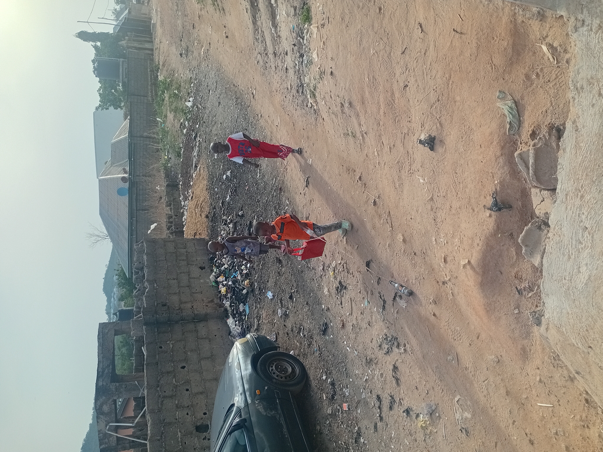 The image depicts a scene of urban decay and apparent poverty in an area identified as Unknown, Unknown. Three children are present, with one wearing a red and white basketball jersey, another wearing an orange shirt, and a third wearing a blue patterned shirt. The children are standing on a dirt path littered with trash and debris. To the left, a dilapidated building with missing sections and crumbling walls is visible, with an old car parked next to it. The ground is a mix of dirt and garbage, with discarded plastic bottles, wrappers, and other waste. In the background, there are simple residential buildings. The sky appears hazy or overcast. The image as a whole conveys an impression of neglect and challenging living conditions.