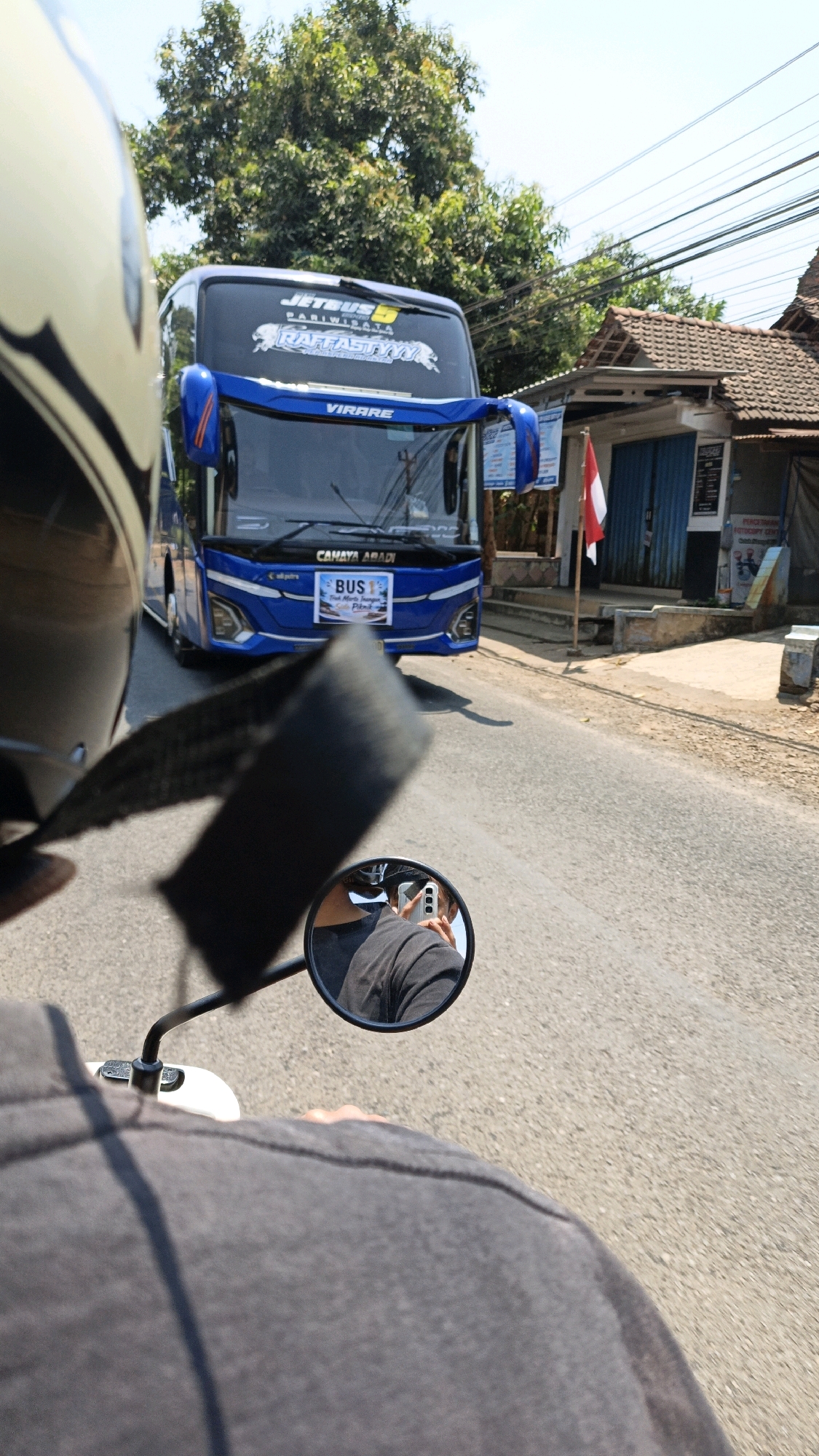 A first-person perspective shot from a motorcycle in Cikal, Indonesia shows a blue double-decker bus ahead. The motorcycle rider, wearing a black t-shirt, is reflected in the side mirror, holding a phone to take a photo. The bus has "JETBUS PARIWISATA" and "RASTAATYYY" written on its rear window, along with a banner that reads "CAHAYA ABADI BUS 1." The bus also has a smaller banner with Indonesian text. The scene is on a paved road with a building and trees on the right side.