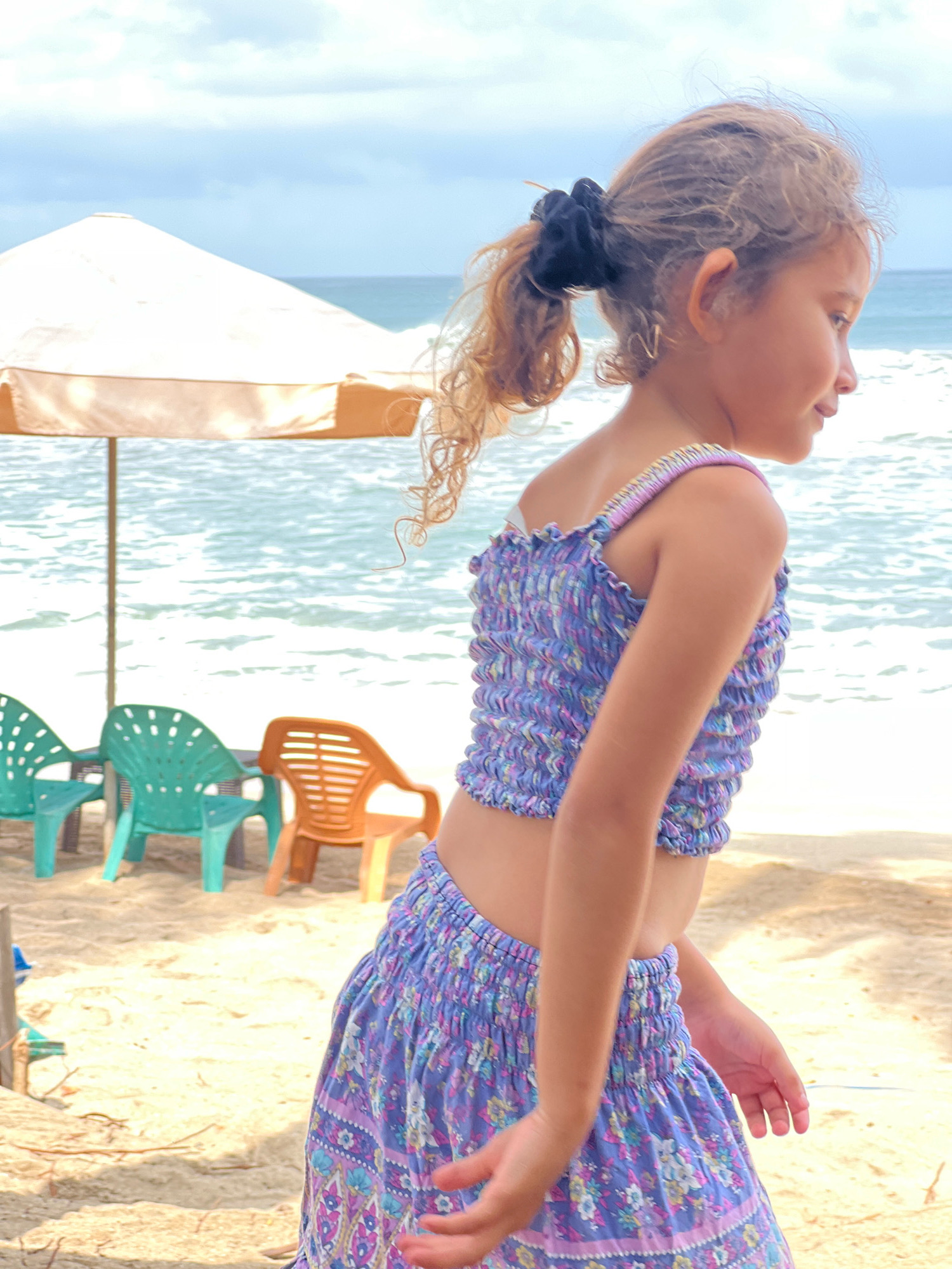 A young girl with curly blonde hair, tied back in a ponytail with a black scrunchie, stands on a sandy beach in Kuta, Indonesia. She is wearing a purple and blue floral patterned two-piece outfit consisting of a smocked crop top and a matching skirt. The girl is facing to her right, looking out towards the ocean.
In the background, the turquoise ocean is visible with small waves. An orange beach umbrella shades two mint green plastic chairs and one orange plastic chair. The sun is bright, indicating it's likely midday. The atmosphere appears relaxed and summery, suggesting a vacation or a day at the beach. No visible text is present in the image.