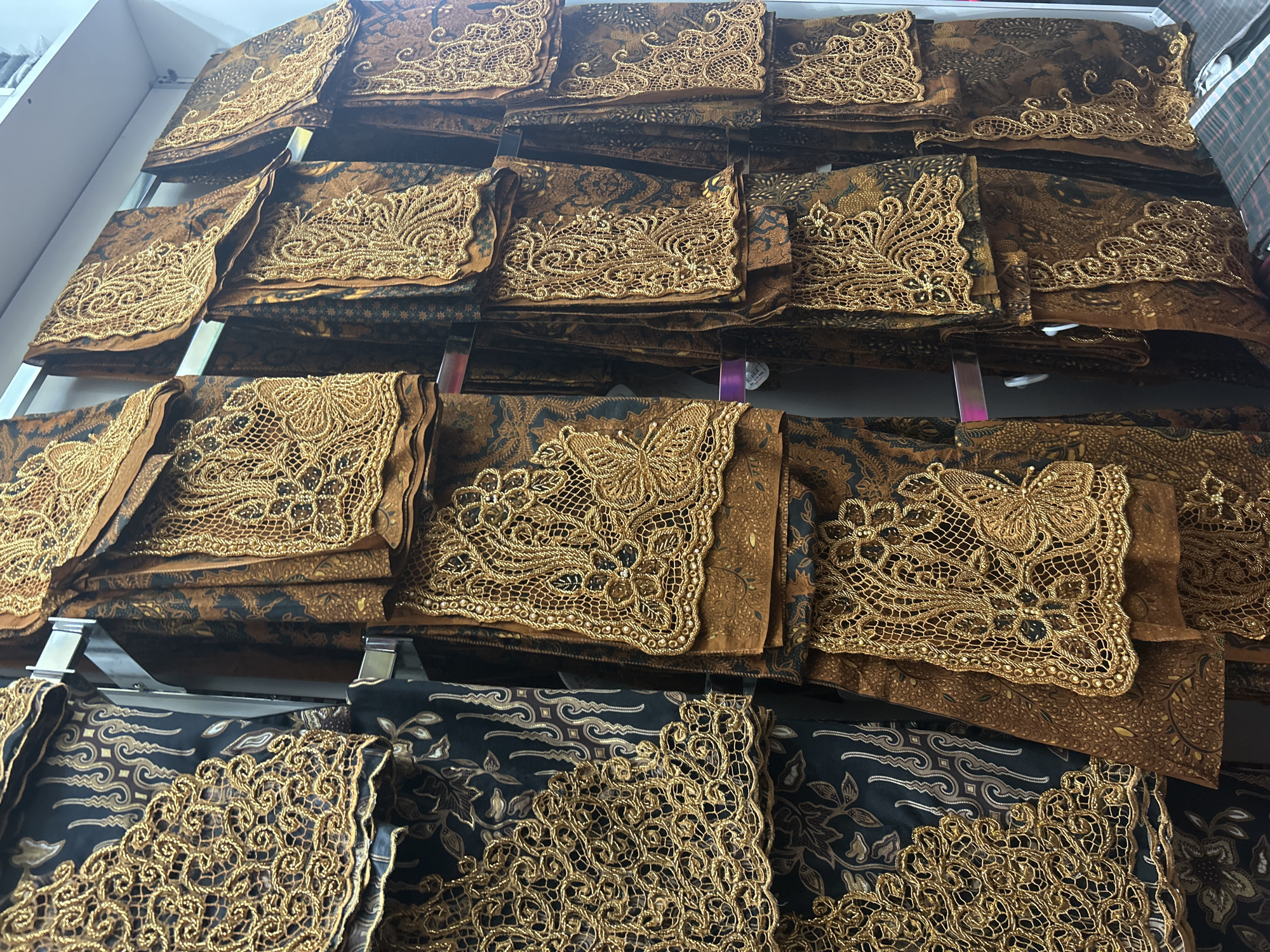 The image shows a retail display of batik fabric with intricate gold lace detailing. The batik patterns are a mix of traditional Indonesian motifs, primarily in brown and gold tones. The lace, also in gold, is applied to the edges and corners of the fabric pieces, featuring floral and butterfly designs.
The fabric is folded and arranged on several shelves or hanging racks, creating a textured and visually rich display. The lighting suggests an indoor setting, likely a shop or market stall. The overall impression is one of traditional craftsmanship and luxury, with the metallic gold lace adding a decorative and opulent element to the fabric. There are no people visible in the image.
The location context of Ubud District, Indonesia, is consistent with the prevalence of batik fabric in the region, known for its artistic traditions. The display appears to be well-organized, showcasing the beauty and detail of each piece of fabric. No visible text is present in the image.