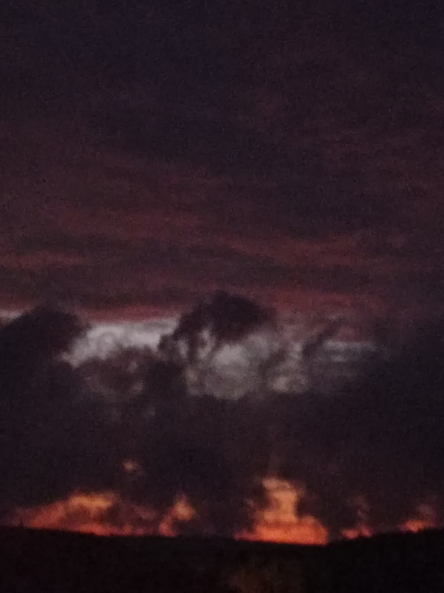 The image shows a dark, cloudy sky at dusk over a silhouetted landscape, likely representing the city of Degtyarsk, Russia. The clouds are predominantly dark purple and black, with hints of red and orange at the horizon, suggesting a sunset or sunrise. The lower portion of the image is obscured by darkness, making it difficult to discern specific details of the terrain or any structures. There are no people or distinct objects visible. The overall atmosphere is somber and muted, typical of twilight conditions.