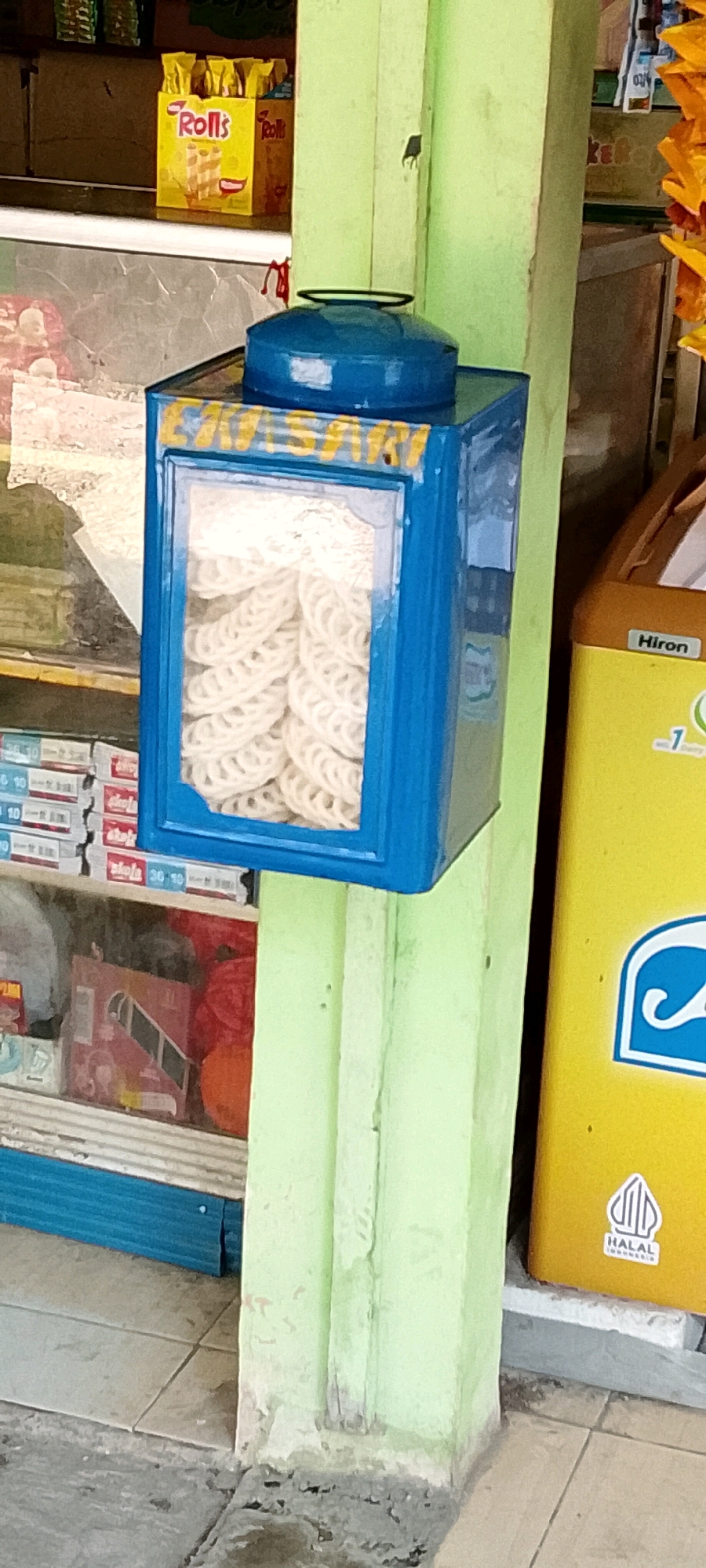 A blue, rectangular display case with a clear window sits on a light green pillar. Inside the case, stacked white, circular snacks are visible. The pillar is located in what appears to be an outdoor market or a small shop in Karangrahayu, Indonesia. To the right of the pillar, a yellow appliance with "Hiron" and a "Halal Indonesia" logo is present. In the background, shelves are stocked with various products, including yellow packaging labeled "Rolls". The ground is tiled.