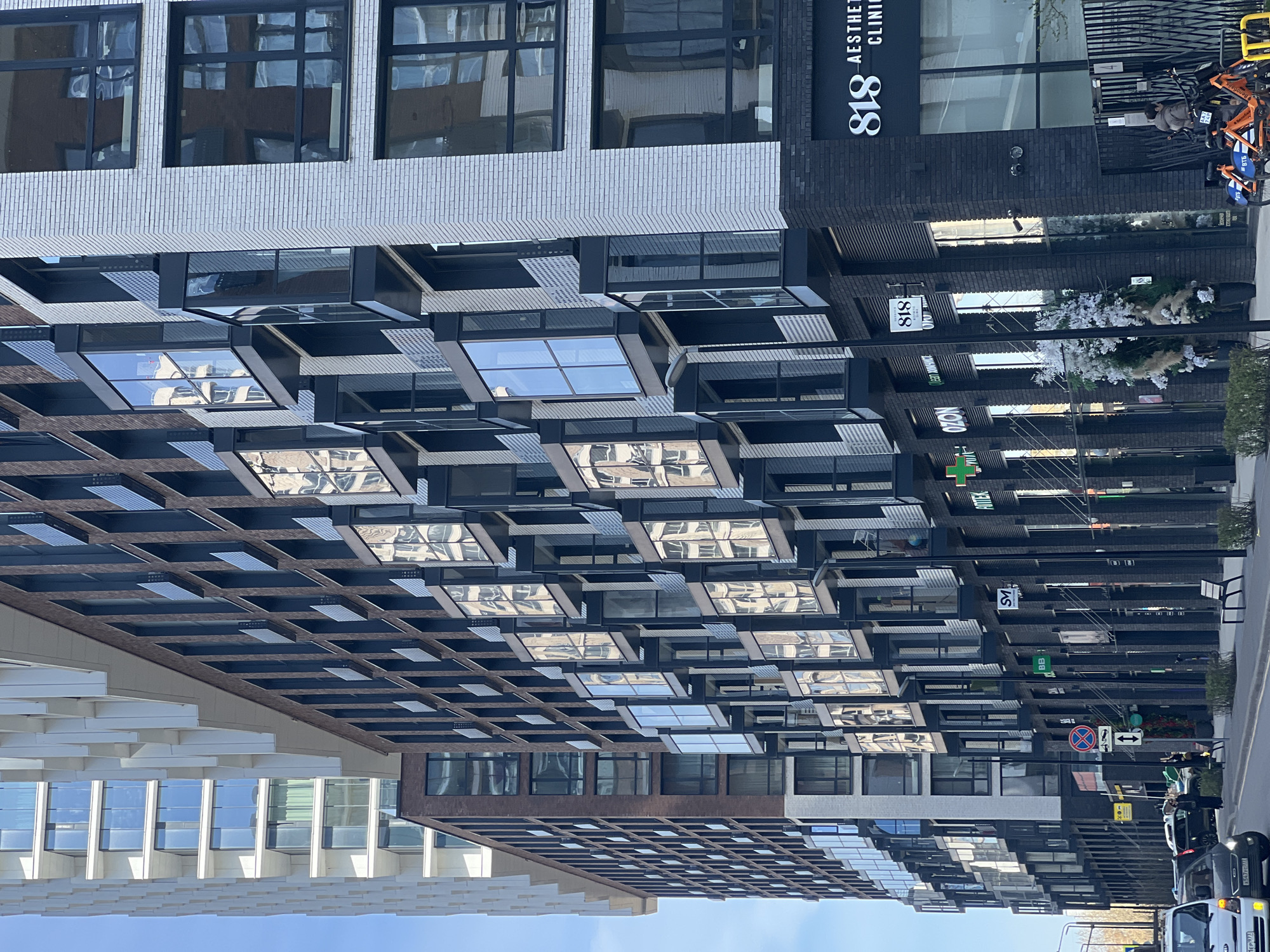 The image captures a modern urban street scene in Khimki, Russia, on a clear, sunny day. Multi-story buildings with contemporary architecture line the street.
The prominent buildings feature varied facades, including light-colored brick on upper levels, dark brick on storefronts, and innovative gridded structures with reflective glass panels that create dynamic reflections of the sky and surrounding architecture. One building on the left shows light brickwork contrasting with a dark, gridded, multi-layered facade beneath, which extends horizontally across the scene.
On the right, a darker brick building houses several commercial establishments. Notable architectural elements include a series of angular, windowed projections extending from the building's facade, offering intriguing reflections.
The street, partially visible on the right side of the image, shows several parked cars, including a white van in the foreground and other vehicles further down the road. Orange bicycles are parked near a storefront, suggesting a bike-sharing service. Street signs, including a 'no stopping' sign and directional arrows, are visible.
Several business signs are displayed on the dark brick facades. Vertically oriented text reads "818 AESTHETIC CLINIC." Other visible signs include a green logo with "OZON," a green cross symbol likely indicating an "APTEKA" (pharmacy), and smaller logos such as "SV" and "BB." White floral decorations adorn the entrance of one establishment. The clear reflections in the glass panels suggest bright daytime conditions. The overall impression is one of a bustling, well-maintained commercial area.