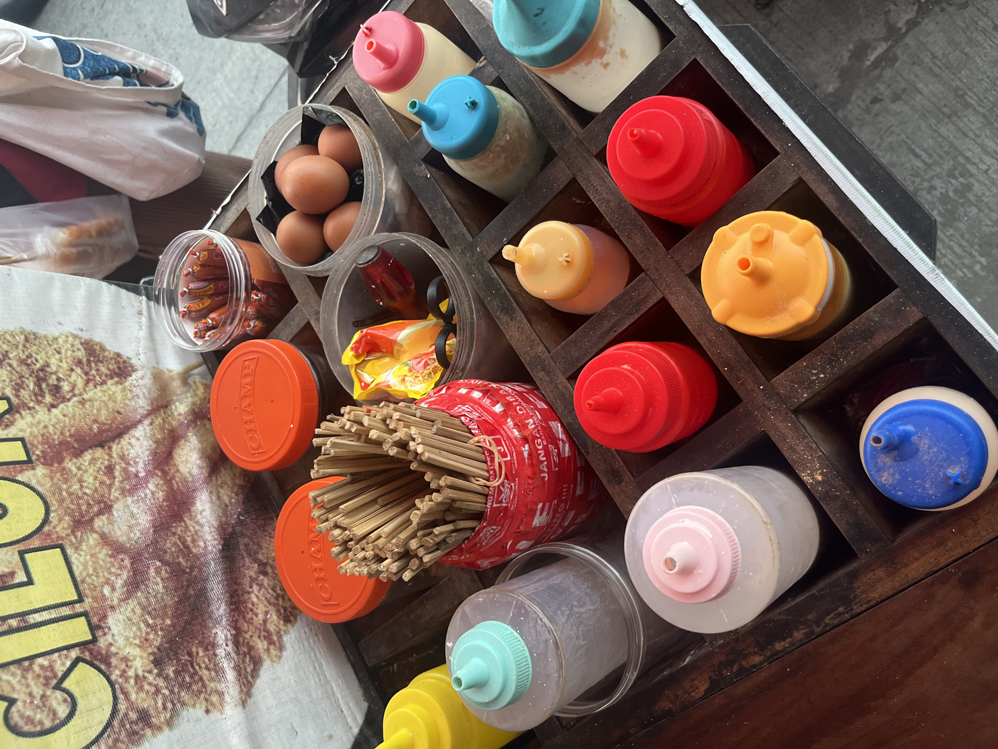The image depicts a close-up, overhead view of a food stall setup, likely for street food. The stall features a wooden organizer with multiple compartments, holding various condiments and food items. Dominating the frame are rows of squeeze bottles with brightly colored caps (red, yellow, blue, pink, and turquoise), some containing sauces and others appearing to be empty or filled with water. In one compartment, a red container holds a large bunch of wooden skewers. Adjacent to this, a clear plastic container is filled with small, individually wrapped chili sauce packets. Further back, a bowl contains several brown chicken eggs. Two orange lids with "CHAMP" branding are visible on top of containers. A large banner with yellow text on a textured brown background is partially visible at the top of the image. The overall setting appears to be outdoors, possibly on a street or sidewalk, with a gray, paved surface in the background. The presence of food items, condiments, and skewers suggests a food preparation or service area. The specific location is identified as Boja, Indonesia.
