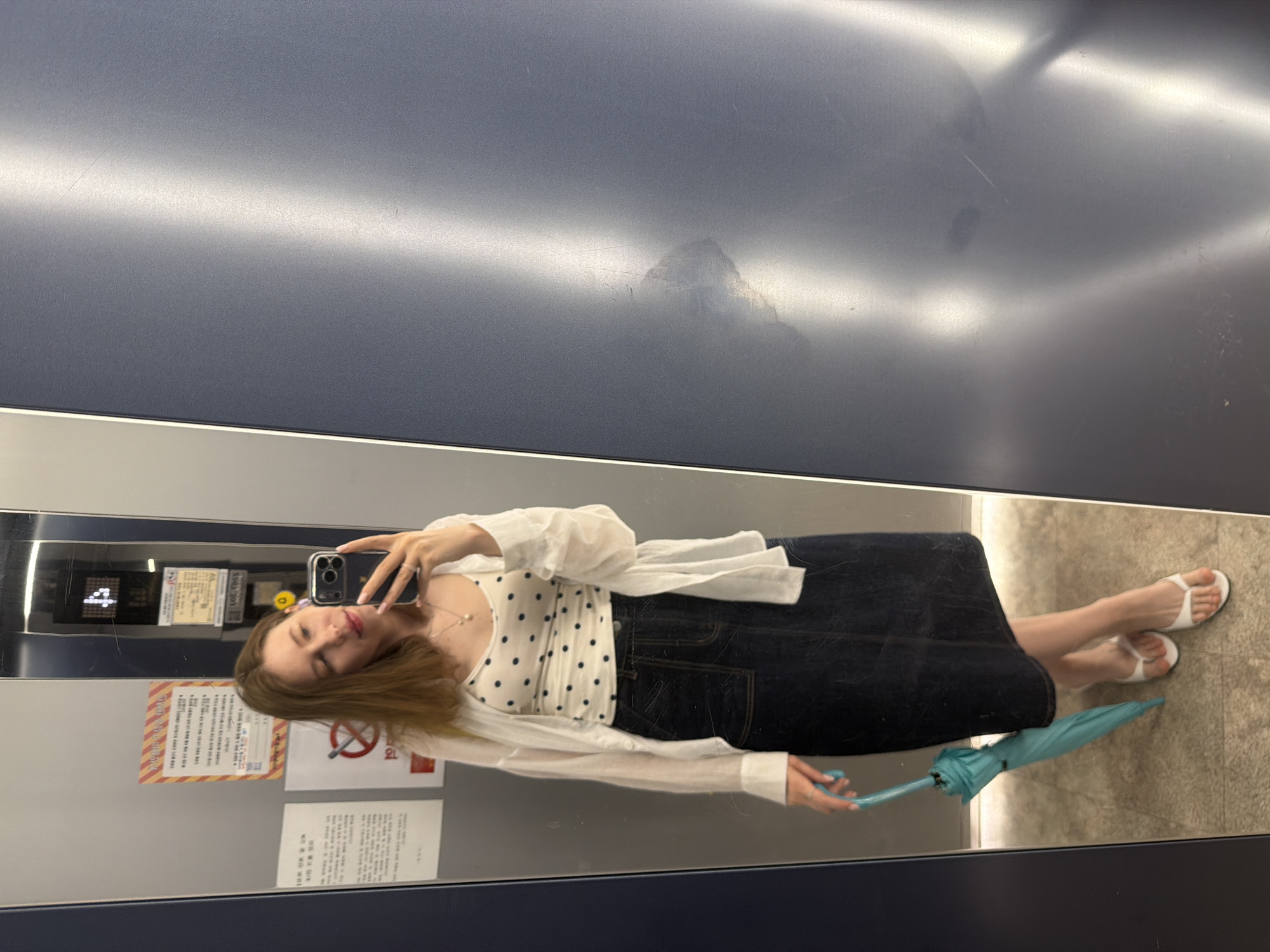 A young woman is taking a mirror selfie in what appears to be an elevator. She is wearing a white polka dot camisole, an unbuttoned white shirt, and a dark denim midi skirt. She is holding a closed turquoise umbrella in her left hand and wearing white thong sandals. She is wearing a necklace with a pearl pendant and a ring on her right hand. In the reflection behind her, there are signs on the wall, one of which has Korean text. The elevator walls are gray and silver. To the right of the elevator doors is a reflective blue surface, possibly another elevator door or wall. The overall environment suggests an indoor, possibly public, space within a building in Dongducheon-si, South Korea.