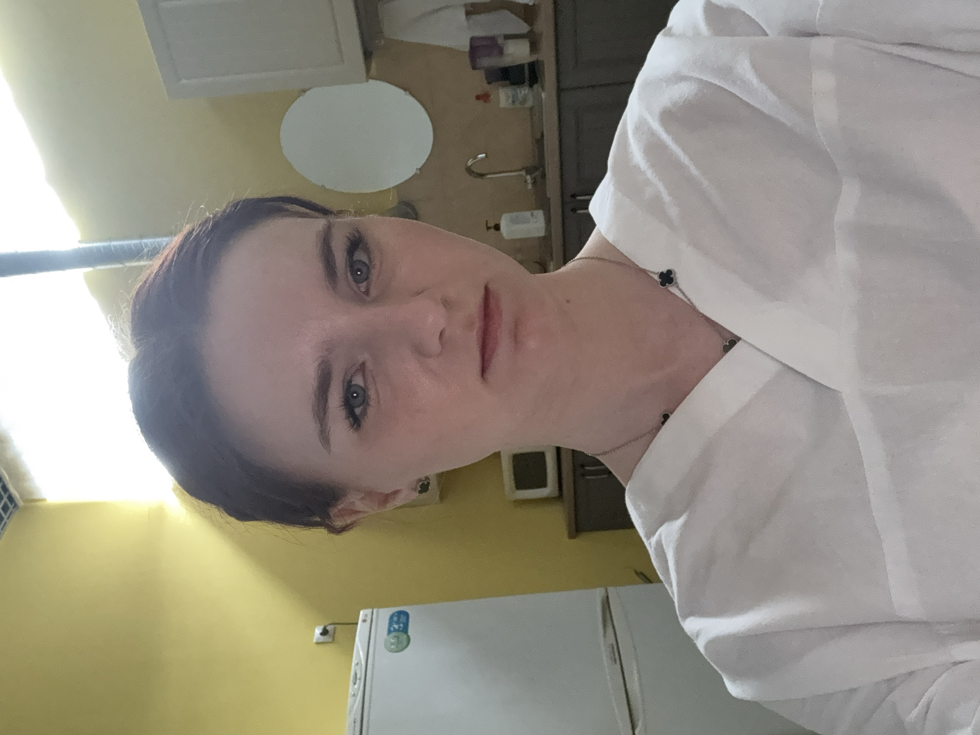 A person is posing for a self-portrait indoors, likely in a kitchen setting. The person appears to be a light-skinned woman with brown hair pulled back, wearing a white blouse and a necklace with a small clover-shaped charm. Her facial expression is neutral.
Behind the person, the background includes a yellow wall, a white refrigerator with magnets, kitchen cabinets, a small round mirror, and a sink with a faucet. There is no conclusive evidence to ascertain the location as Saint Petersburg, Russia. The environment is a typical domestic kitchen.