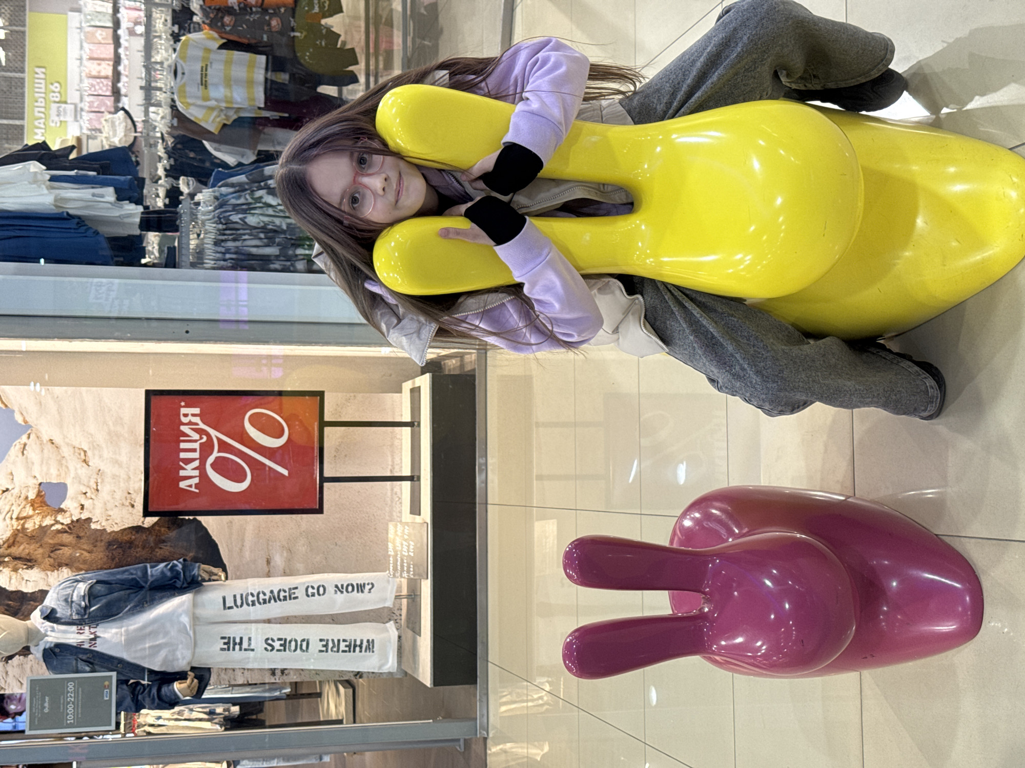 A young girl wearing eyeglasses, a light purple hooded jacket, gray pants, and black gloves is lying on a yellow, decorative, bunny-shaped object in an indoor shopping mall setting. She is smiling at the camera while embracing the yellow object. To the right of her is another similar decorative piece that is pink. Behind her is a retail storefront displaying clothing and mannequins. Visible within the storefront's window is a promotional poster with "АКЦИЯ" (translated to "SALE") and a percentage sign in Russian, as well as signs reading "WHERE DOES THE LUGGAGE GO NOW?" The mannequins display blue denim jackets and white pants with text. The image is taken from a high-angle, slightly tilted perspective.