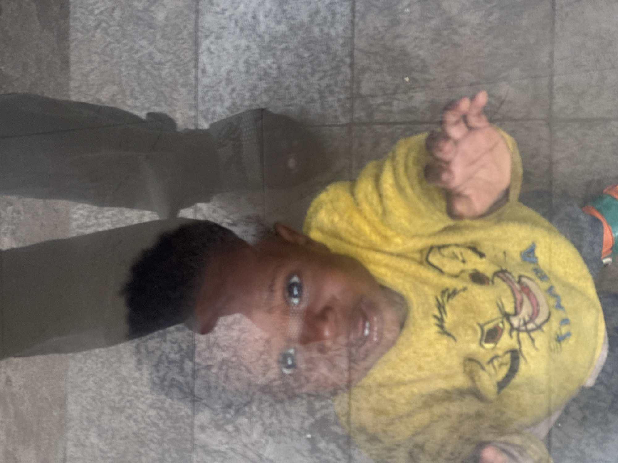 The media file captures a close-up, low-angle shot of a young child wearing a yellow t-shirt with a graphic of a cartoon character, possibly Simba from The Lion King, printed on it. The child is looking up and to the right, with wide eyes and a slight smile. Their right hand is raised and slightly blurred, suggesting movement or a gesture. The child's skin tone is dark, and they have dark hair.
The background appears to be a textured, tiled surface, possibly a floor or pavement. The lighting is somewhat diffused, and there are reflections on the surface, indicating it might be wet or covered in a reflective material. The overall scene is indoors or in a covered outdoor area, given the absence of strong natural light or weather elements.
There is some faint, blue text on the t-shirt, partially obscured by the graphic. The text is difficult to decipher due to its blurriness and the design elements of the shirt.
The image is presented in a portrait orientation, but the subject is oriented such that the child's head is towards the bottom of the frame and their feet are towards the top, suggesting the photo was taken from a high vantage point looking down. The setting does not provide specific cues about the time of day or weather beyond the potential wetness of the ground. The child's expression is one of curiosity and engagement.