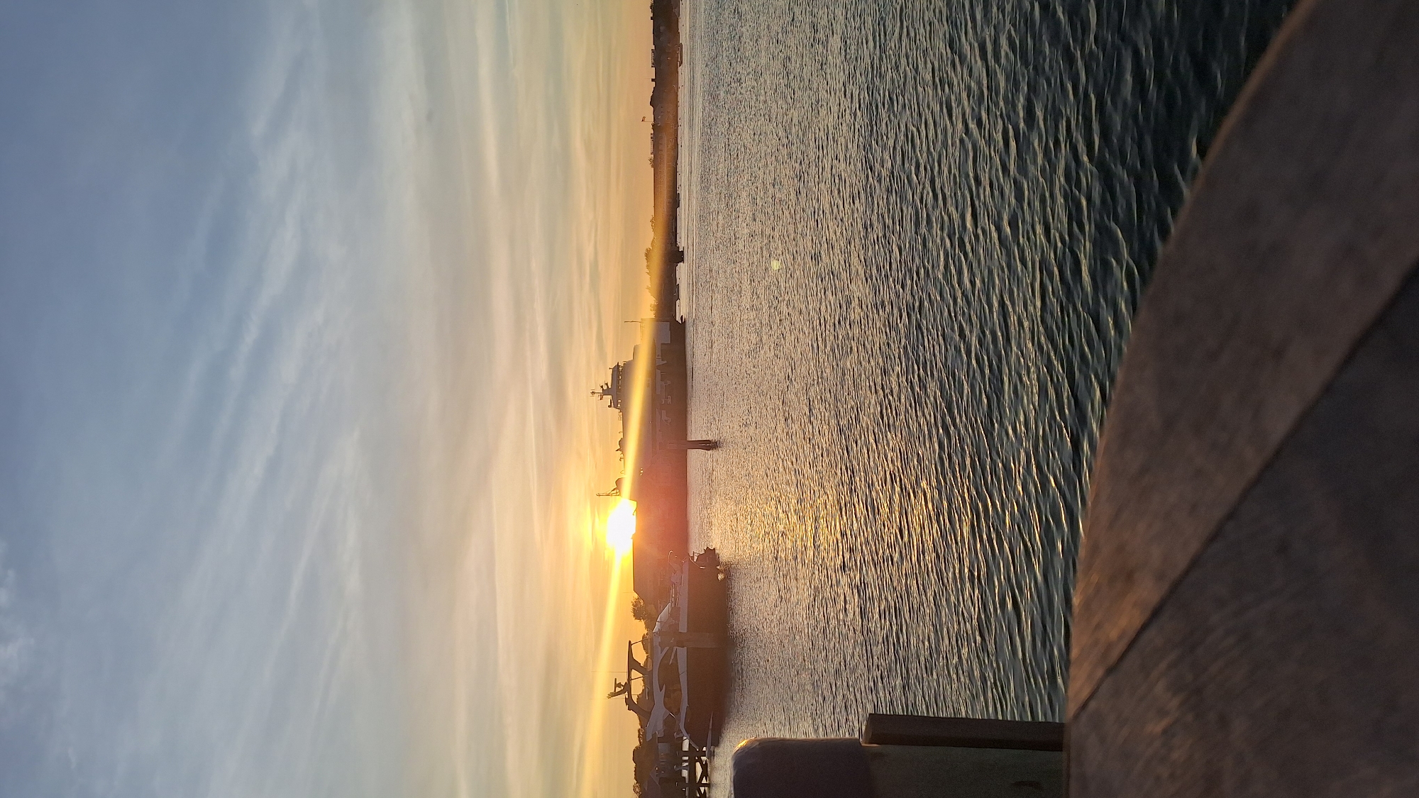 This media file captures a tranquil harbor scene during either sunrise or sunset, likely in Nantucket, United States of America. The image is oriented horizontally, showcasing the sun low on the horizon, casting a brilliant golden light across the sky and water.
The sky gradients from a soft blue on the left to vibrant oranges and yellows near the sun on the right. Wispy, elongated clouds stretch across the sky, illuminated by the warm light. The water in the foreground and mid-ground reflects the golden hues of the sun, creating a shimmering, rippled surface.
Several boats and structures are silhouetted against the bright horizon. A larger vessel, possibly a ferry or cargo ship, is visible in the mid-distance. Closer to the viewer, smaller boats appear to be docked or moored. A distant landmass or shoreline runs across the horizon, providing a backdrop to the maritime activity. In the lower right foreground, a wooden railing or pier structure extends diagonally into the frame, indicating the photograph was taken from a dock or elevated position.
The time of day is clearly dawn or dusk, characterized by the intense, low-angled sunlight. The water appears calm with gentle ripples, and the sky is mostly clear with light clouds, suggesting pleasant weather. The overall mood conveyed is peaceful and serene. No specific human activity or visible text is present in the image.
