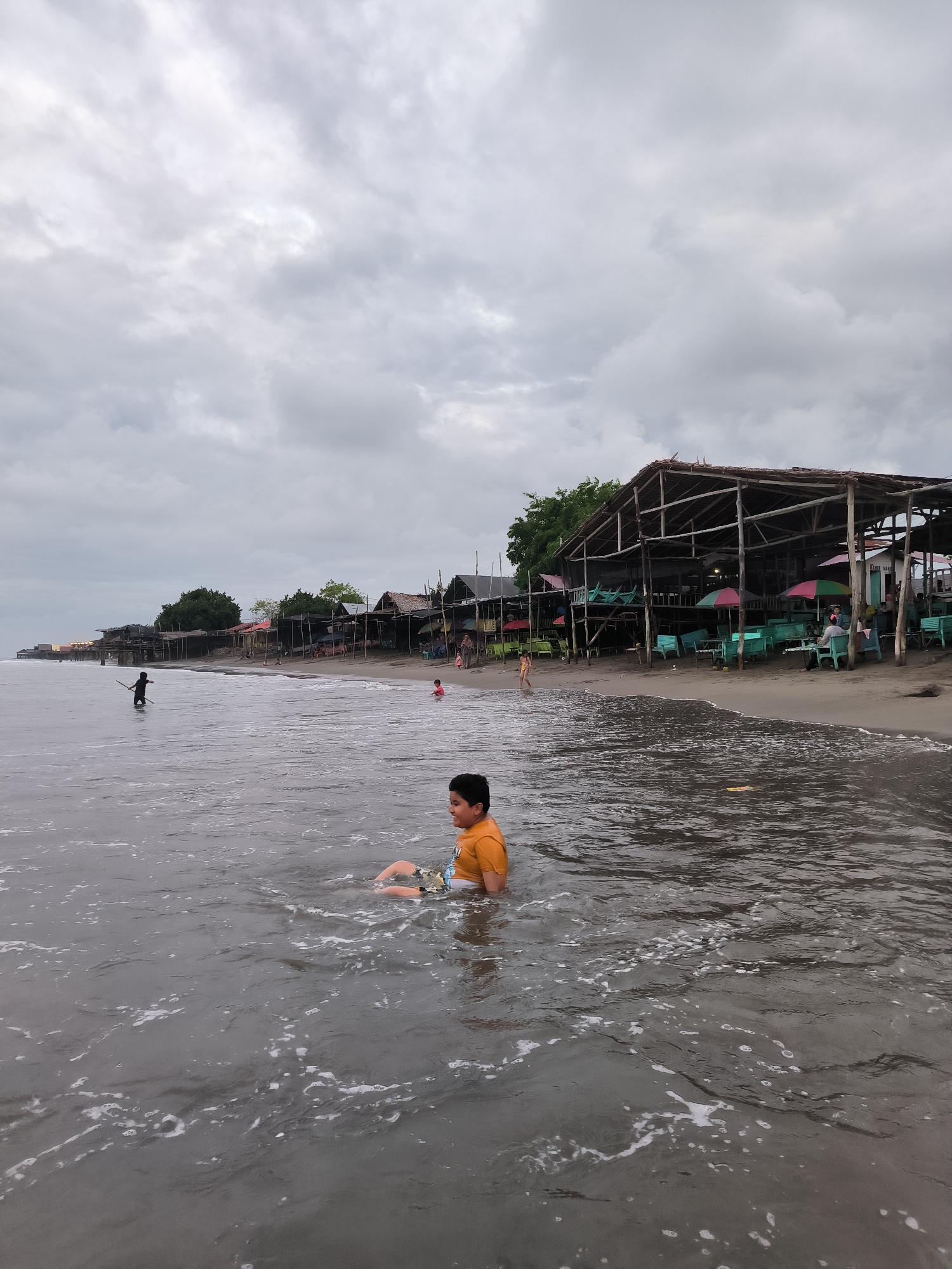 The image depicts an overcast day at a coastal beach in Lhokseumawe, Indonesia. In the immediate foreground, dark, rippled shallow sea water with white foam from small waves extends across the frame. A young boy, wearing an orange t-shirt and patterned shorts, is seated or crouched in the water on the right side, facing left. Further back in the water, a child in dark clothing holds a stick-like object while standing, and another child in pink clothing is visible near the shore on the right.
The midground features a dark sand beach lined with numerous rustic, open-air structures, primarily constructed from wood with thatched or corrugated iron roofs. Many of these structures contain green or teal colored tables and chairs, with some featuring colorful umbrellas (pink, green, red). People are visible sitting at these tables, walking on the beach, or standing near the structures. Behind the structures, coastal vegetation, including green trees, is present. The coastline stretches into the distance on the left, revealing additional structures. A muted grey, cloud-filled sky dominates the upper portion of the image.