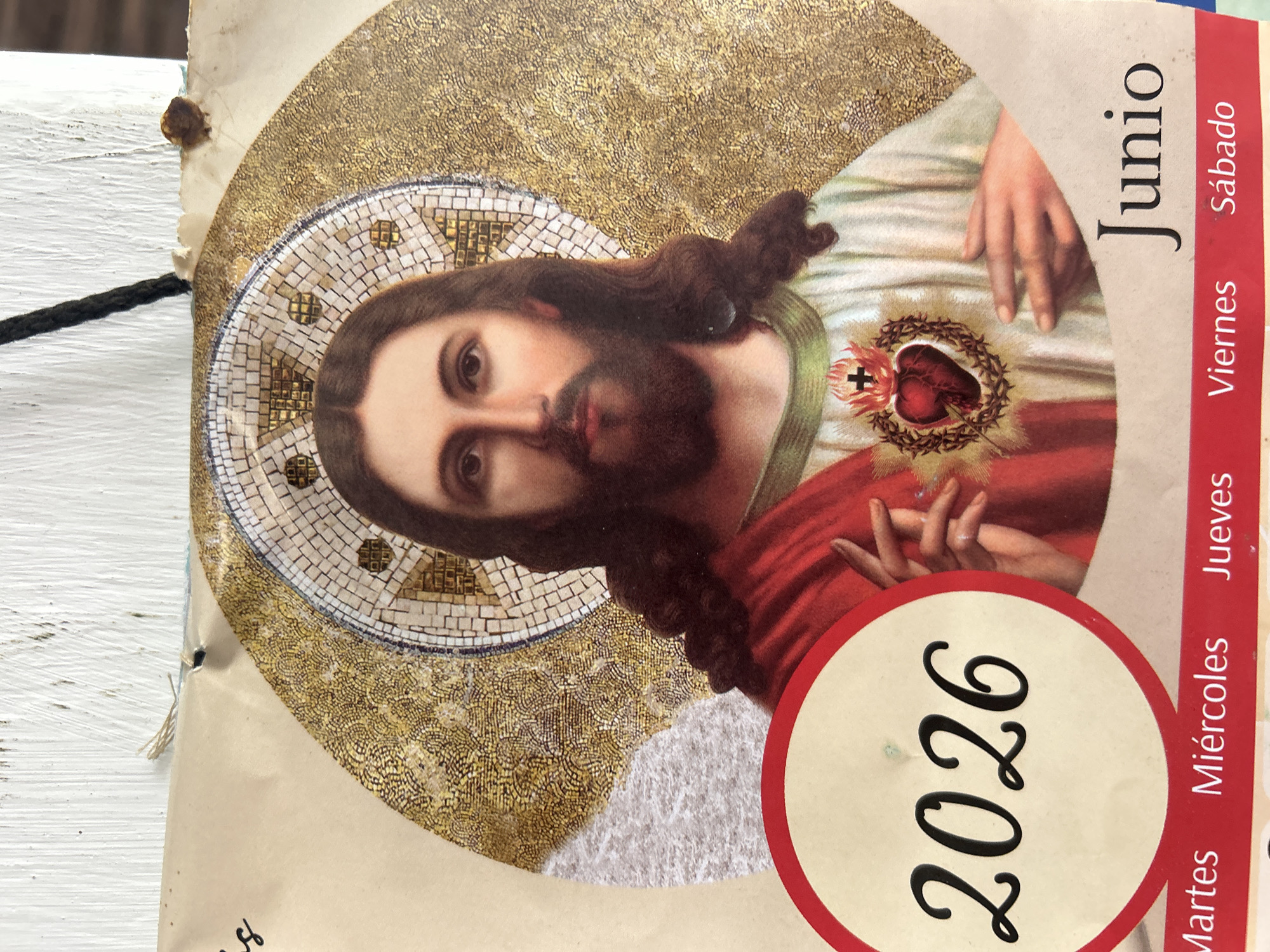 The image is a close-up of a calendar featuring a religious illustration of Jesus Christ. He is depicted with a beard, long curly brown hair, and a halo. His chest bears the Sacred Heart, enclosed in a thorny crown, with a cross on top. He is wearing a red robe over a white garment. The background is adorned with a mosaic-style halo and a gold, textured pattern.
In the lower left quadrant, a circular emblem contains the year "2026" in large, decorative black numerals. Below this, along the bottom of the image, is a red band with the days of the week printed in white: "Martes", "Miércoles", "Jueves", "Viernes", and "Sábado". To the right of the year emblem and above the day names, the word "Junio" is printed in large, black, serif font.
The calendar appears to be attached to a textured, light-colored surface, possibly a wall or a wooden plank, with a dark string hanging from the top. There are small imperfections and tears visible on the edge of the calendar paper.
While the image is from a calendar, there is no information within the image itself that indicates its specific location in Santa Ana, Venezuela, or any other city.
