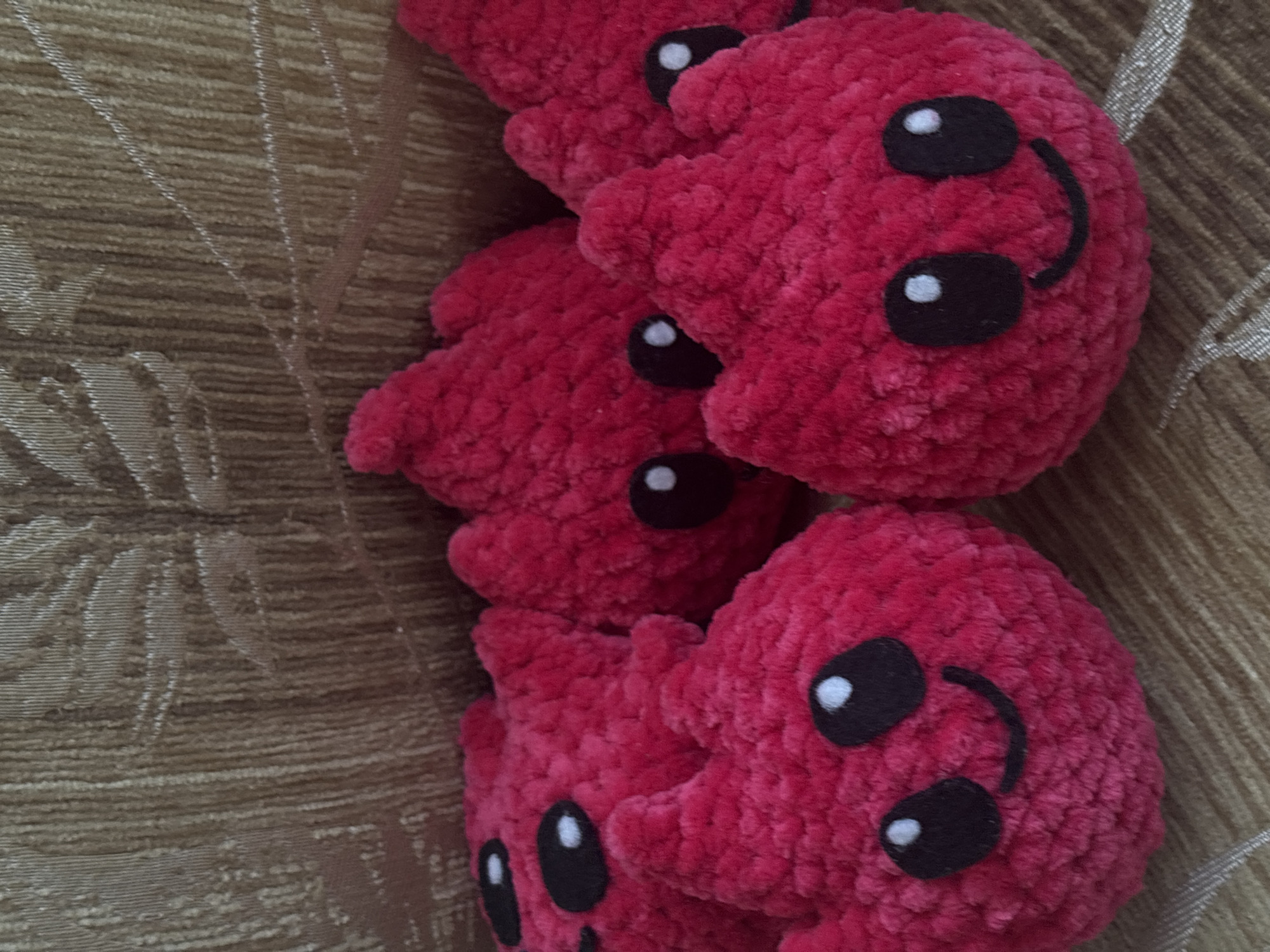 This is a close-up, overhead, eye-level shot of five crocheted, fuchsia-colored, heart-shaped objects with embroidered black eyes and smiling mouths. They are arranged in a cluster on a textured, golden-brown fabric. The objects are soft and appear to be plush toys or decorative items. The fabric background has a raised, floral pattern. The overall scene is well-lit, and the focus is sharp on the crocheted objects. There is no indication of a specific location in Varna, Bulgaria.