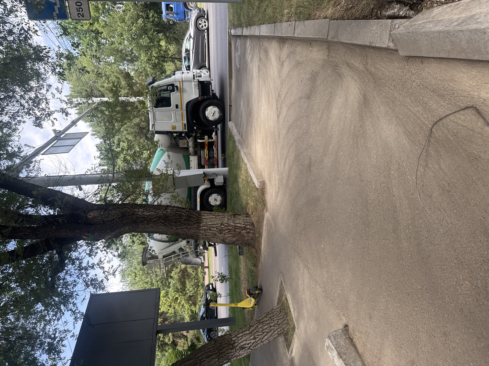 A yellow electric scooter is parked on a concrete path next to a tree. In the background, a white cement mixer truck is stopped on a road, with a dark-colored car behind it. A blue truck is visible further down the road. The scene is set in a city with trees and foliage, under a bright sky with some clouds. A traffic sign indicating "250" is also visible. The location is on the street near the Almaty Arbat, a pedestrian zone in Almaty, Kazakhstan.
