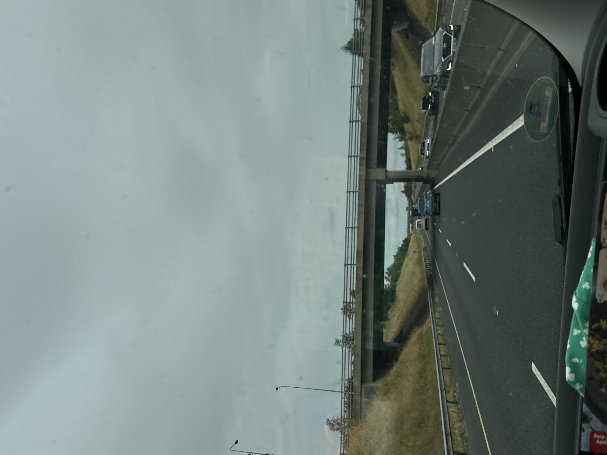 The image is taken from inside a vehicle, looking forward on a multi-lane highway under a cloudy sky. A concrete overpass stretches across the highway. On the overpass, there are railings and some vegetation on top. On the sides of the highway, there are grassy embankments and guardrails. Several cars are visible on the highway, traveling in the same direction as the camera's vehicle. One car, a dark SUV, is in the right lane. Another car, a light-colored sedan, is in the center lane, and a blue car is in the left lane. The scene appears to be within the vicinity of Moone, Ireland, as indicated by context. The road surface is asphalt, with white lane markings. The overall environment is that of a typical highway setting.