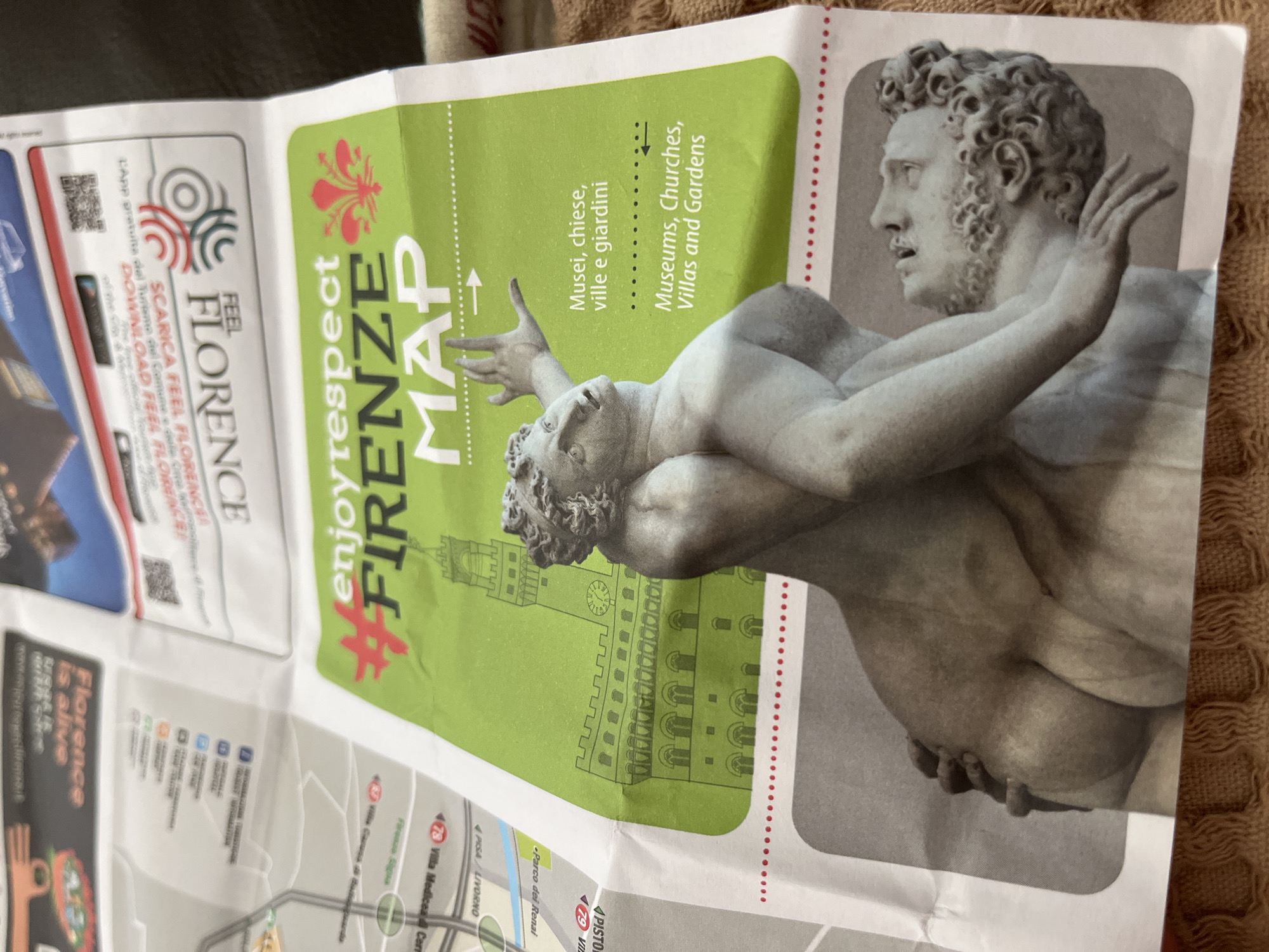 A folded tourist map or brochure for Florence, Italy, lies open and angled on a textured brown fabric surface. The central image on the brochure features a large, grayscale depiction of a classical marble sculpture, showing two muscular figures intertwined. One figure, with curly hair and a beard, looks upwards and to the right with raised arms, while the other is partially visible beneath.
The top portion of the brochure has a light green banner with red and white text. Prominently displayed is a red fleur-de-lis, the symbol of Florence. Large text reads "#enjoyrespect FIRENZE MAP." Below the fleur-de-lis, smaller text provides categories for tourist attractions in both Italian and English: "Musei, chiese, ville e giardini" and "Museums, Churches, Villas and Gardens." An outline illustration of a historic building, possibly Palazzo Vecchio, is visible behind the sculpture on the green banner.
To the left, a white and red section encourages app downloads with text like "FEEL FLORENCE," "SCARICA FEEL FLORENCE" (Download Feel Florence), and "L'APP gratuita del Turismo del Comune di Firenze" (The free app of the Municipality of Florence Tourism). A small black and red box in the bottom left corner states "Florence is alive." Parts of an actual map are visible at the bottom, with some legible place names such as "Parco dei Renai," "PISA / LIVORNO," and "Villa Medicea Cerr," indicating locations around Florence. The map's details are somewhat blurry, while the sculpture and banner text are clearer.