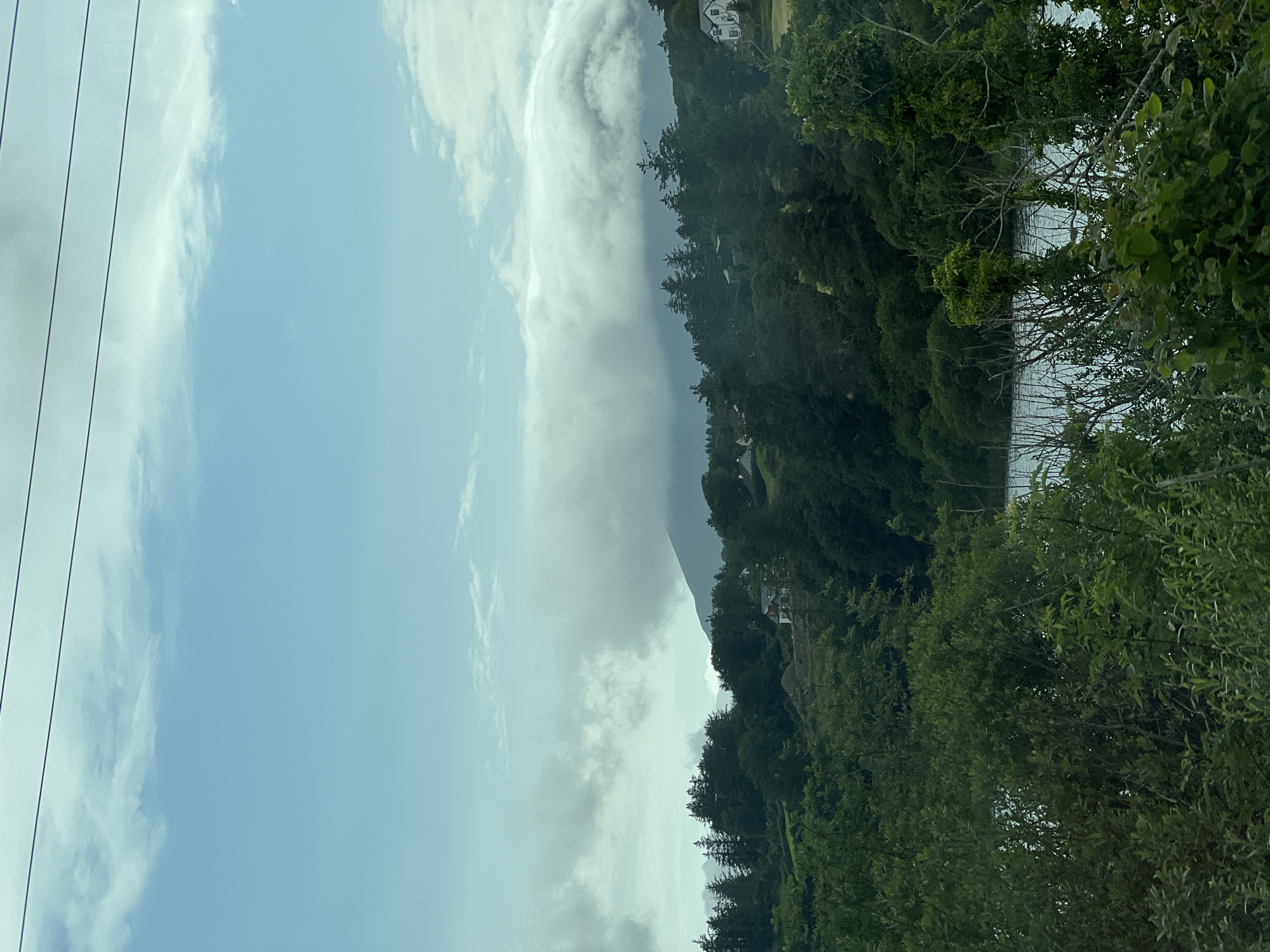 The image shows a scenic view of a cloudy sky over a body of water, with lush green trees and vegetation lining the shore. The perspective is from a high vantage point, looking down towards the water and the wooded hillside. There are a few houses visible nestled amongst the trees on the slope. The sky is partly cloudy with dramatic formations of white and grey clouds, suggesting a dynamic weather pattern. The time of day appears to be daytime, possibly late afternoon or early evening, given the soft lighting. The location is identified as Creeslough, Ireland, which is consistent with the verdant, coastal landscape. The overall mood is serene and natural, highlighting the beauty of the Irish countryside. There are no visible people or objects that suggest specific activity, but the presence of houses indicates a residential area within this natural setting. The image evokes a sense of tranquility and the raw beauty of nature.