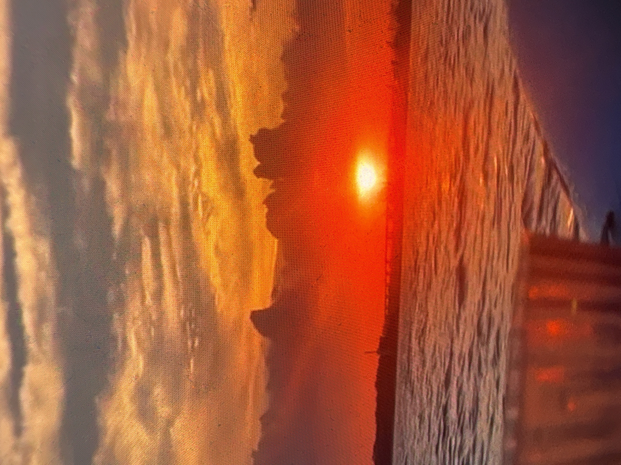The image depicts a sunset with the sun appearing as a bright white spot, positioned near the center of the frame. The scene has warm hues of orange and yellow, with some areas appearing darker. The image is out of focus, with the pixels showing. There are suggestions of buildings present on the right side of the image. The location is unidentified.