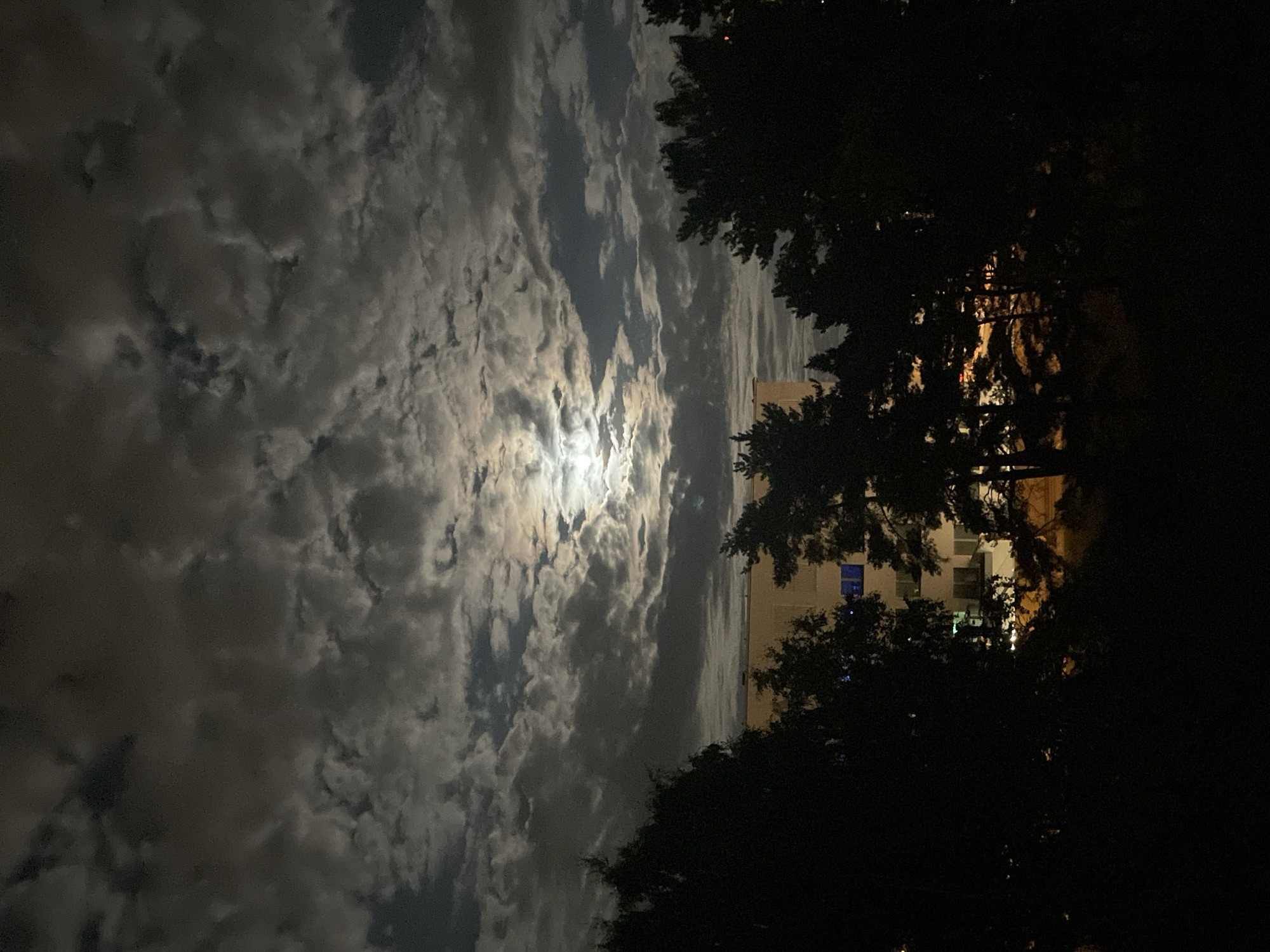 This image shows a cloudy night sky with the moon visible through a gap in the clouds. In the foreground, silhouetted trees obscure a building. The building appears to be an apartment block with some illuminated windows, suggesting it is in a residential area. The image was taken at night, as indicated by the darkness and artificial lighting. The presence of a building and trees suggests an urban or suburban environment. The image does not provide enough detail to determine the specific location within Tallinn, Estonia.