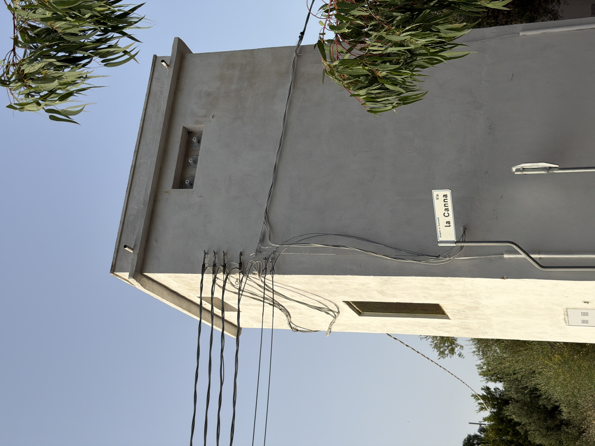 The image shows a light gray, rectangular, multi-story structure, possibly a utility building or watchtower, in Santu Diadòru/San Teodoro, Italy. The building has a flat roof with a short parapet and a small pipe protruding from the top. A rectangular window with a metal grate is visible on the left side of the building. Several electrical wires run horizontally across the upper left portion of the image and are attached to the building with various brackets and conduits.
On the front of the building, a white street sign is mounted on a gray pole. The sign reads "Burgeta la Suaredda Via la Canna". Below the window, a white electrical box with a grated cover is mounted on the wall.
The building is situated outdoors, with a clear blue sky in the background. Lush green foliage from a tree with long, narrow leaves hangs into the frame from the upper right and lower right portions of the image. The foliage appears to be eucalyptus. In the distance, to the left, there are more trees and vegetation, suggesting a rural or semi-rural setting within Santu Diadòru/San Teodoro. The lighting suggests it is daytime, possibly late afternoon, given the shadows. No people are visible in the image.