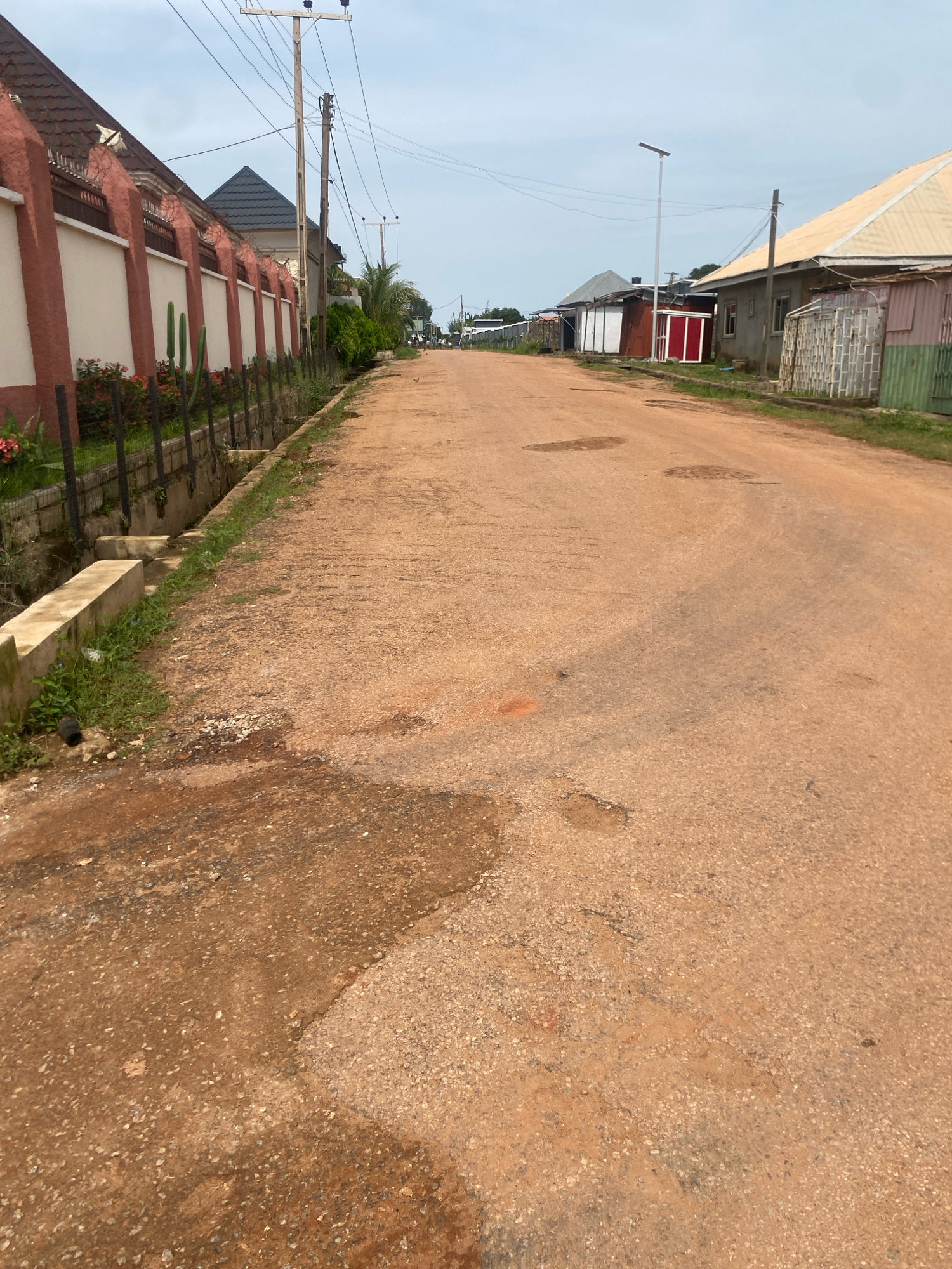 The image depicts a street scene in Barden Yana, Nigeria. The unpaved road, composed of reddish-brown earth and gravel, shows signs of wear and tear with several noticeable potholes and uneven patches. The road is bordered on the left by a low concrete drainage channel and a fence made of concrete posts and metal mesh, behind which a residential property with a red and white wall and a dark tiled roof is visible. Beyond this property, more residential structures with metal roofs and some vegetation are seen. On the right side of the road, several single-story buildings with corrugated metal roofs and varying facade materials are present, including some with what appear to be metal sheeting and others with plastered walls. A solar-powered street light stands tall on the right side of the road in the middle distance. Overhead, a network of electrical wires is strung between utility poles. The sky above is overcast with a pale, diffused light, suggesting a cloudy day. The overall impression is that of a residential area with modest infrastructure.