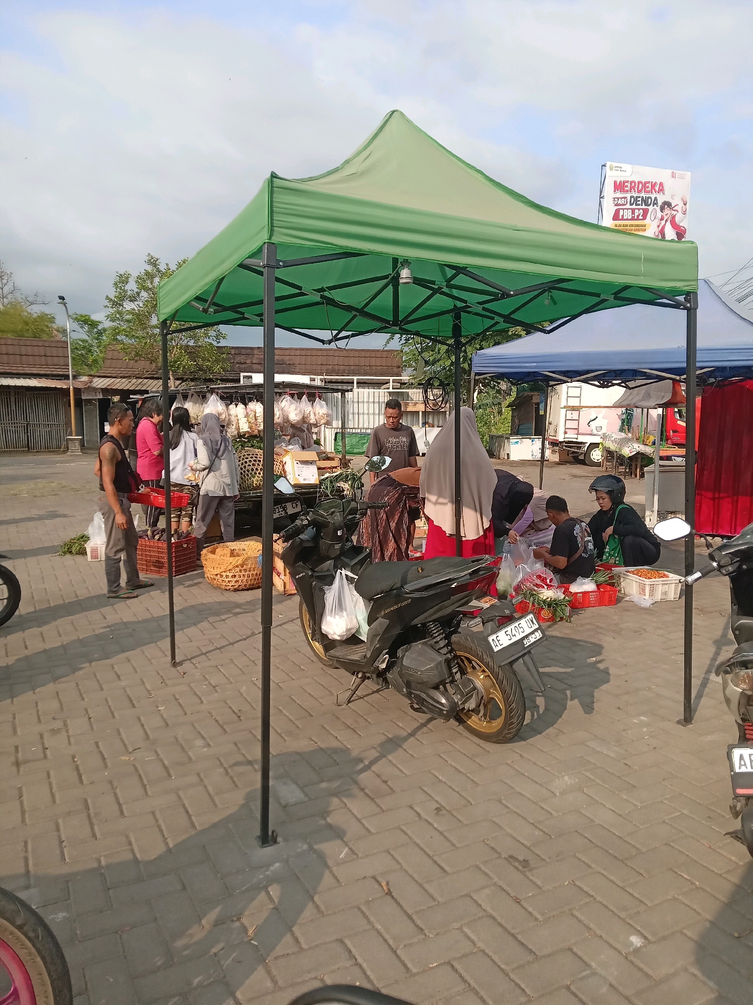This image depicts an outdoor market scene in Banguntapan, Indonesia. A green canopy tent provides shade over a stall where vendors and customers interact. Several people are present, some appearing to be selling goods and others browsing. Various items are displayed, including what appear to be fruits, vegetables, and packaged goods, some contained in woven baskets and red crates. Two motorcycles are parked near the stall, one in the foreground and another partially visible to the right. The ground is paved with interlocking bricks, and shadows suggest it is daytime with the sun casting light on the scene. In the background, there are buildings with tiled roofs, trees, and a billboard displaying promotional content. The overall atmosphere suggests a typical market day.