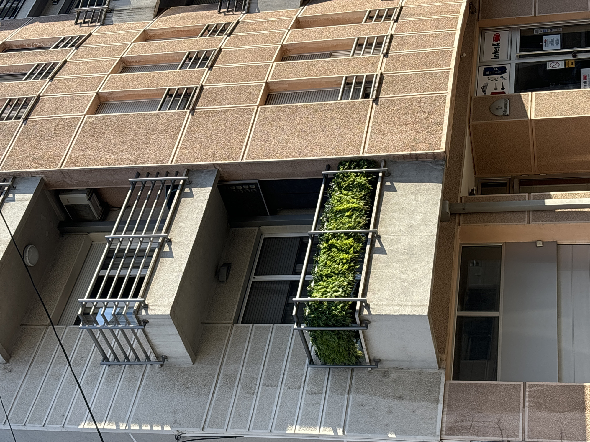 A low-angle, eye-level exterior shot captures a segment of a multi-story building in Vracar Urban Municipality, Serbia. The building's facade exhibits a mix of textures and colors. The left side of the building features a light-colored, textured concrete wall with horizontal grooving. On this section, a balcony with a metal railing and a trough of lush green plants is visible. Above this balcony, another balcony with a similar metal railing is seen. The right side of the building is characterized by rectangular panels of a reddish-brown, textured material separated by white trim. This section has multiple windows with metal shutters.
Below the balconies and windows, the ground level of the building reveals a business establishment with a glass storefront. Signage on the storefront includes "Intesa" and various icons. The entrance appears to be a commercial door. A metal garage door is also visible on the far left side of the image, at the base of the building. The lighting suggests it is daytime, with shadows cast across the building's facade. The overall scene depicts an urban architectural environment.