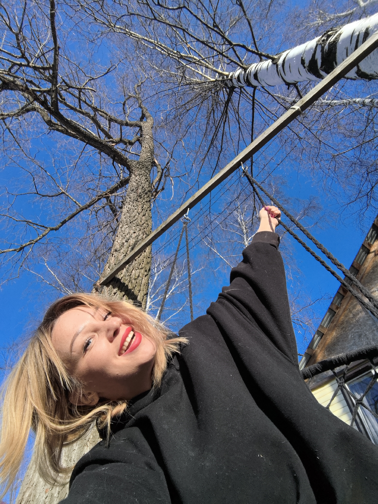 A woman with short blonde hair, bright red lipstick, and a black hoodie is smiling and looking up in Noviy, Russia. She is holding onto a rope that is attached to a metal bar suspended between two trees, a birch and a dark-barked tree. A building with yellow siding is partially visible in the background against a clear blue sky. The trees are mostly bare, with only a few branches.