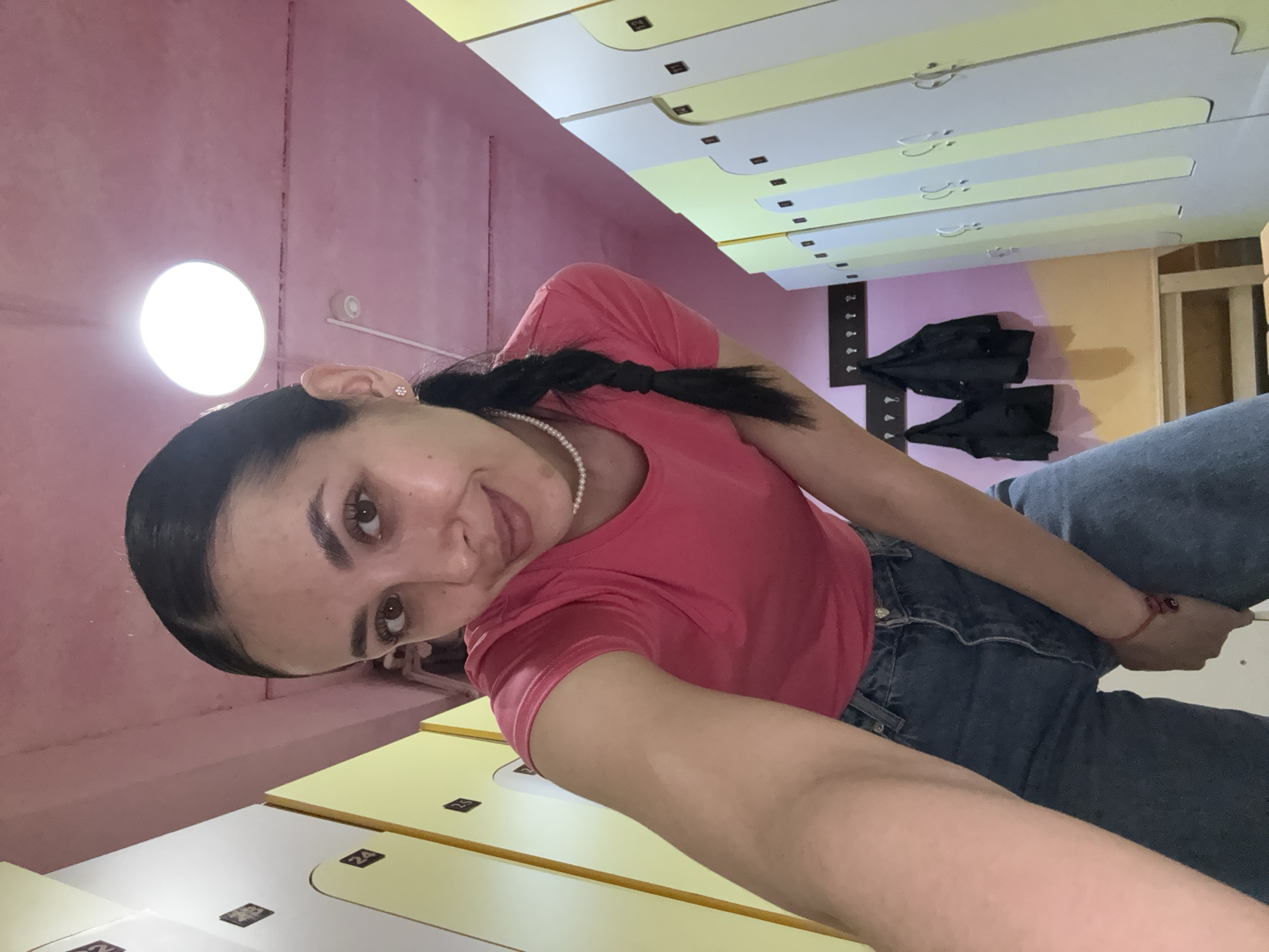 The image shows a woman with dark hair styled in a braid and wearing a pink t-shirt, a pearl necklace, and jeans. She appears to be in a locker room, as indicated by the rows of lockers in the background. The lockers are yellow and white, and the walls are painted pink and yellow. There is a coat rack with dark jackets hanging on it in the background. The room is lit by a bright, round light fixture. There is no information in the image to suggest this location is Votkinsk, Russia.