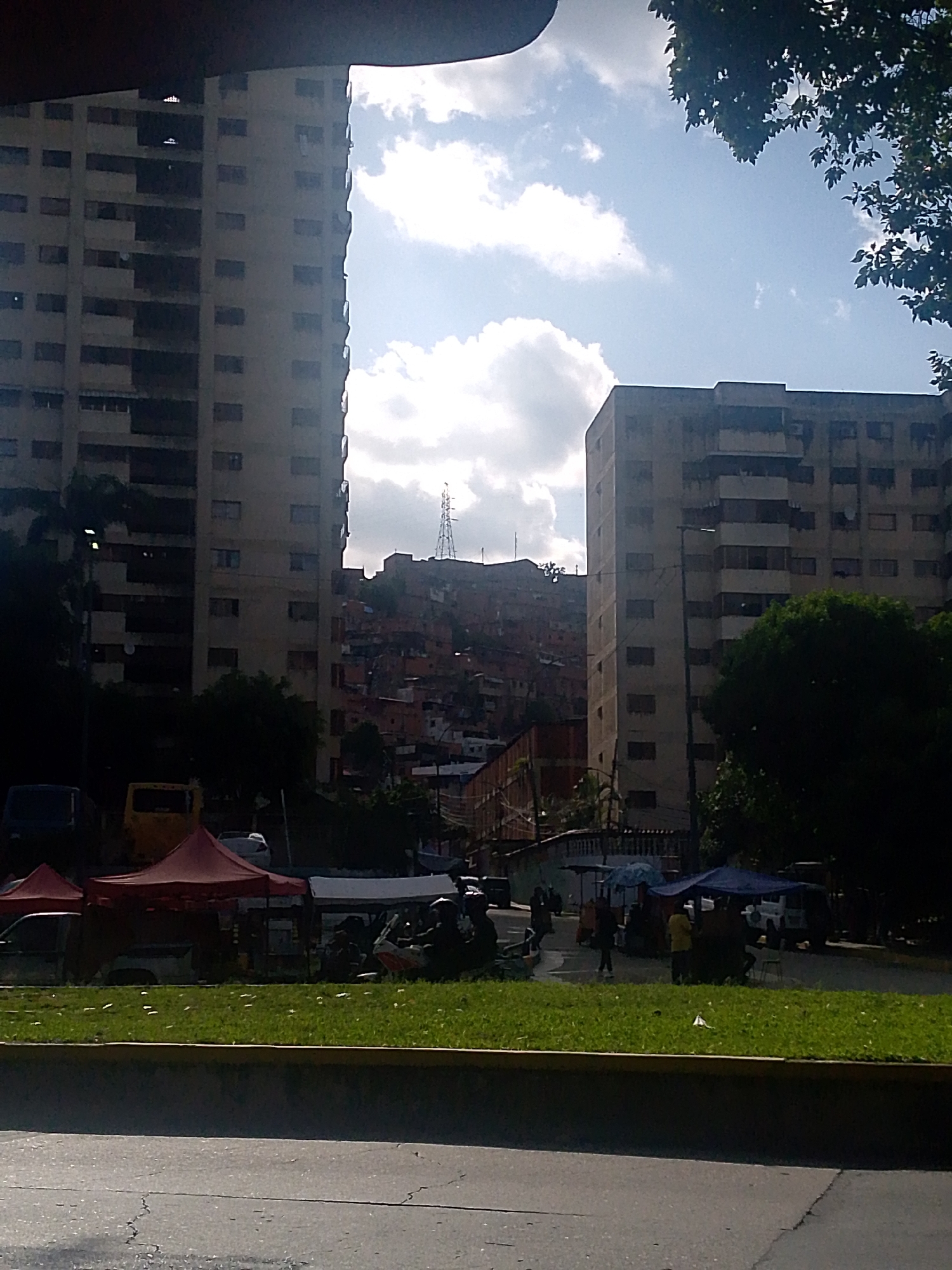 The image captures a view of a hillside favela in Caracas, Venezuela, with two large apartment buildings in the foreground. The favela, characterized by densely packed, colorful houses with corrugated metal roofs, sprawls across the elevated terrain. A prominent cell tower is visible on the highest point of the hill, indicating its importance as a communication hub.
In the foreground, the scene transitions to a more urban setting with a grassy median strip separating the road from a sidewalk area. Several market stalls are set up under colorful tents, with vendors and shoppers engaging in commerce. Red tents dominate the left side of the frame, while white and blue tents are visible further to the right. A yellow bus is parked behind the red tents, and various cars are also present. The overall impression is one of a vibrant, bustling urban environment, showcasing the stark contrast between formal apartment buildings and the informal settlements on the hillside, a common characteristic of Caracas. The lighting suggests it is daytime, with the sun illuminating the sky and casting shadows.