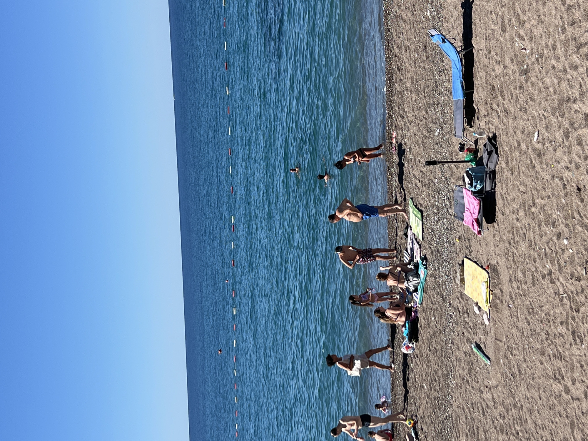 This image depicts a sunny day at a pebble beach in Rafailovići, Montenegro. The clear blue Adriatic Sea stretches towards the horizon, marked by a line of buoys. On the shore, several people are enjoying the beach. Some are in the water, swimming and wading, while others are on the sand. A group of people are gathered near the water's edge, some appearing to be talking. Several beach chairs, towels, and bags are scattered on the sand, indicating people are relaxing and sunbathing. The overall atmosphere is one of leisure and summer vacation.