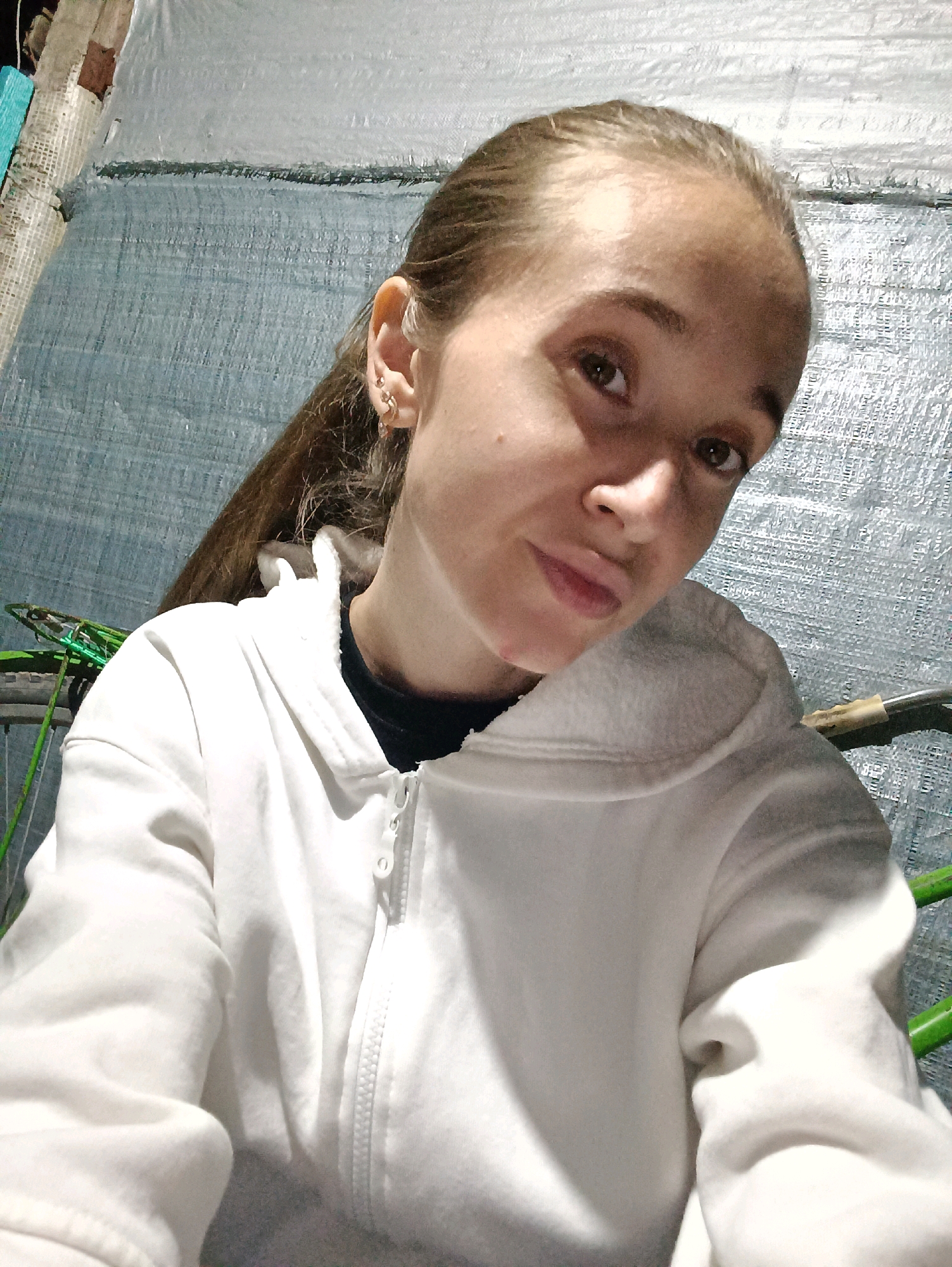 A young woman with light brown hair pulled back in a ponytail is taking a selfie. She is wearing a white zip-up hoodie over a dark shirt. She has multiple earrings in her left ear. She is indoors, with a textured blue tarp as the background. Green metal bars are visible to the left and right of the frame. The lighting suggests it is evening or night. No specific location in Vypasne, Ukraine, can be determined from the image.