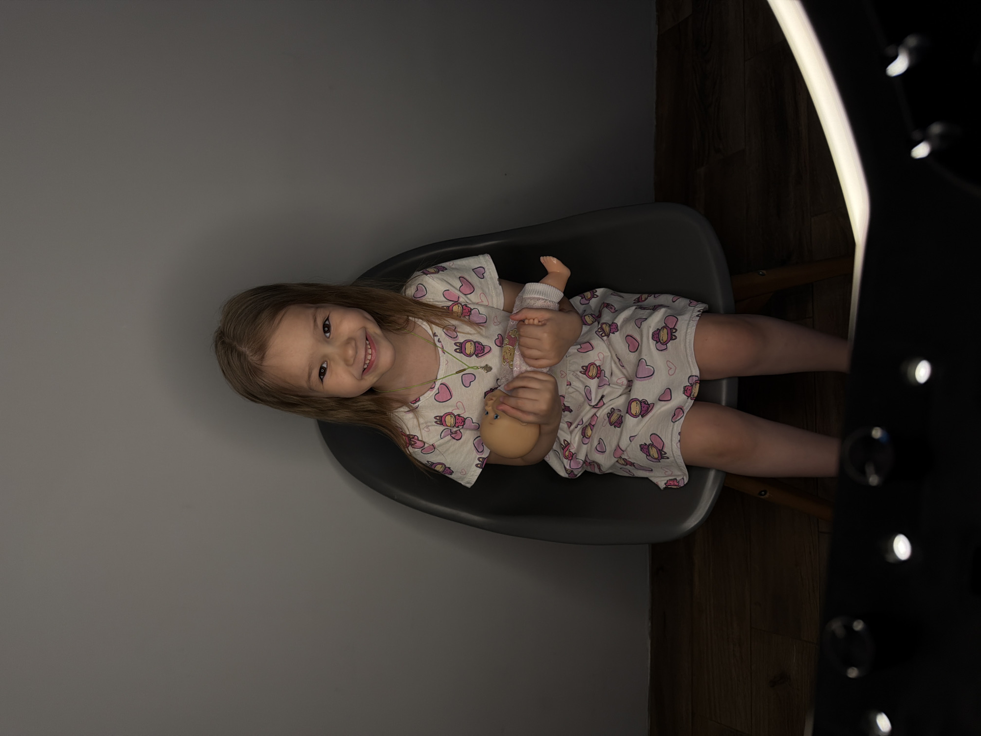 A young girl with blonde hair sits in a gray chair, holding a baby doll. She smiles towards the viewer. The scene appears to be indoors, with a gray wall and wooden flooring. A ring light fixture is visible on the right. There is no specific information in the image to verify the location as Chaikovsky, Russia.