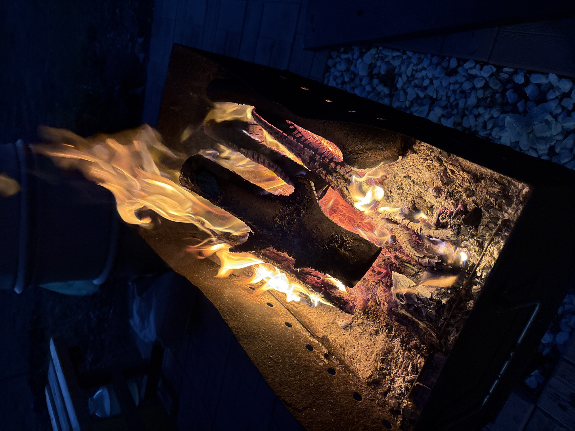 The image shows a square metal fire pit filled with burning logs. Flames are visible, particularly on the left side of the pit. Ash and embers are present within the pit. The surrounding environment appears to be a patio or outdoor area with wood decking and small decorative stones visible near the edge of the fire pit. The scene is lit primarily by the fire itself, with the background appearing dark. The location is not discernible as Dubovaya, Russia. There is no evidence of any people or interactions in this image.